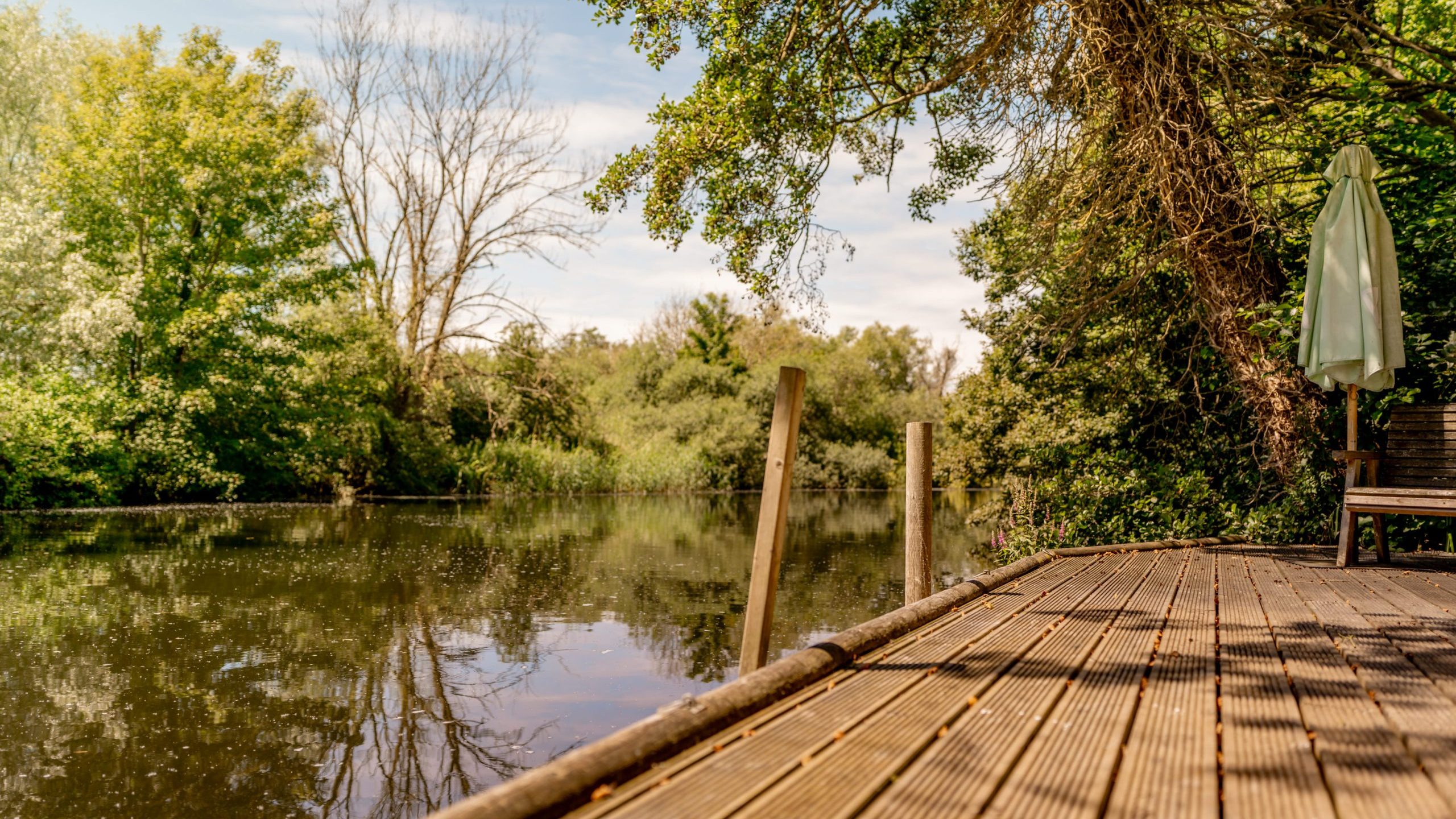 A wooden dock extends over a calm river surrounded by lush green trees under a partly cloudy sky. A bench and closed umbrella sit on the dock’s edge, creating a peaceful, shaded spot by the water.