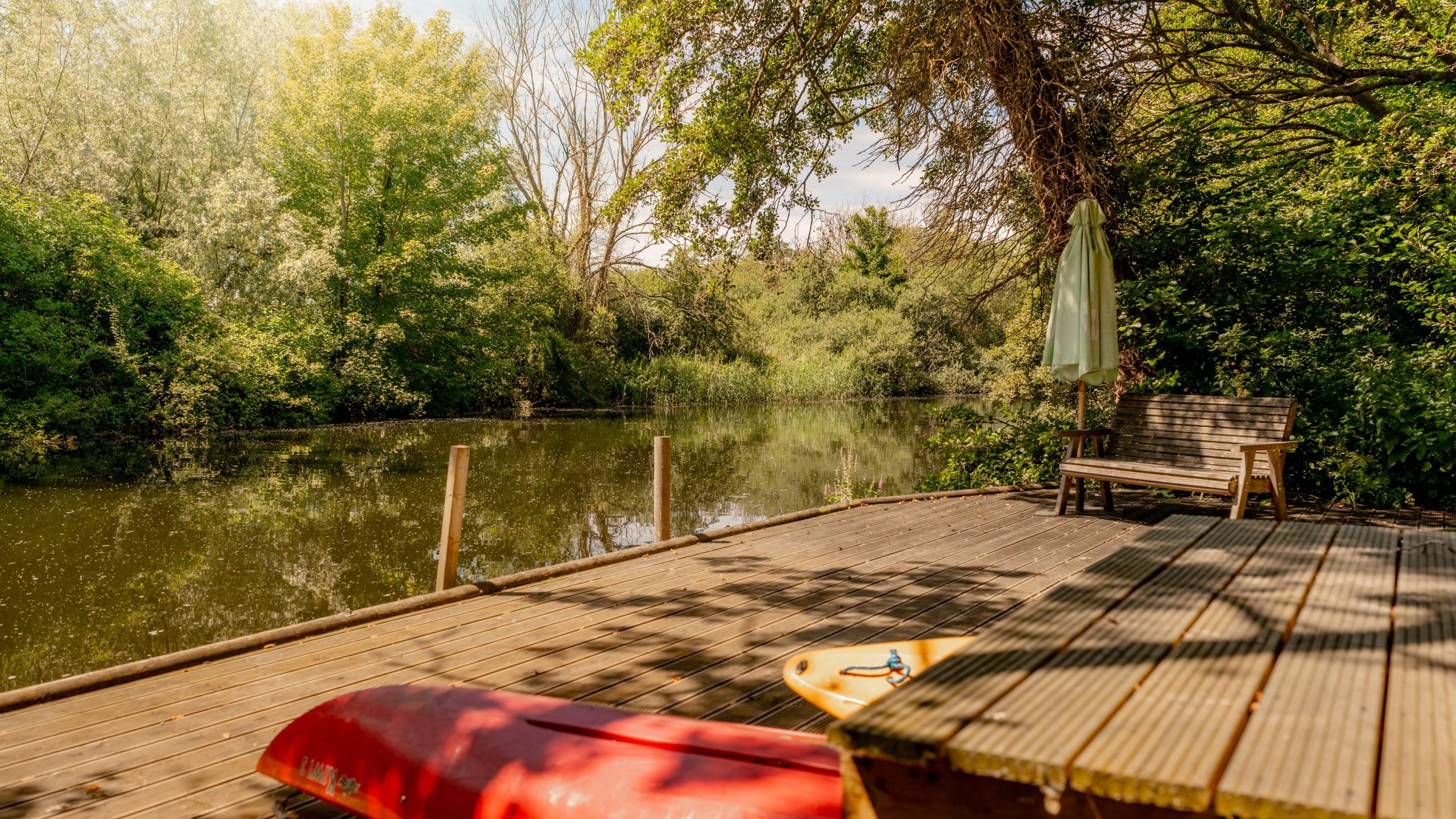 A wooden deck beside a calm river is surrounded by lush green trees. A red canoe, paddle, bench, and closed umbrella are on the deck under dappled sunlight.