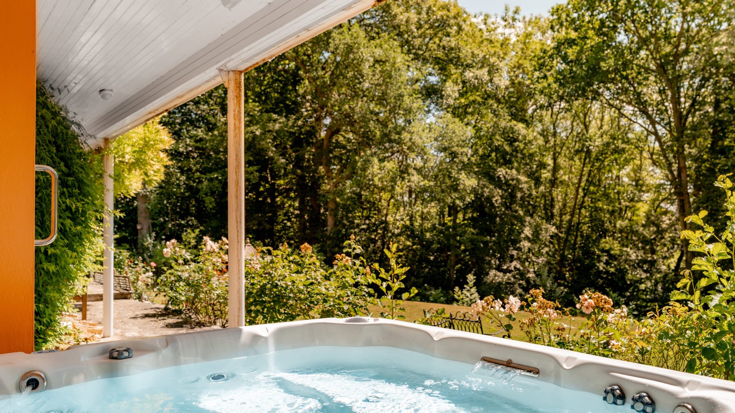 An outdoor hot tub with bubbling water under a covered patio, surrounded by greenery, flowering plants, and tall trees on a sunny day.