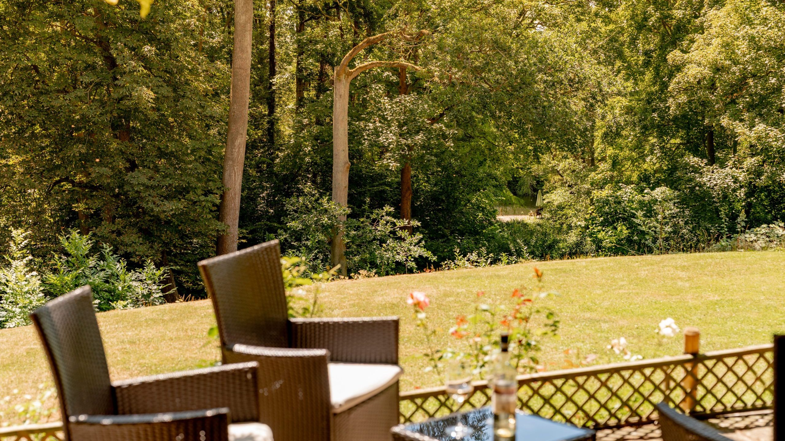 Wicker chairs and a small table with flowers sit on a patio overlooking a sunlit grassy lawn and a lush, green forest in the background.