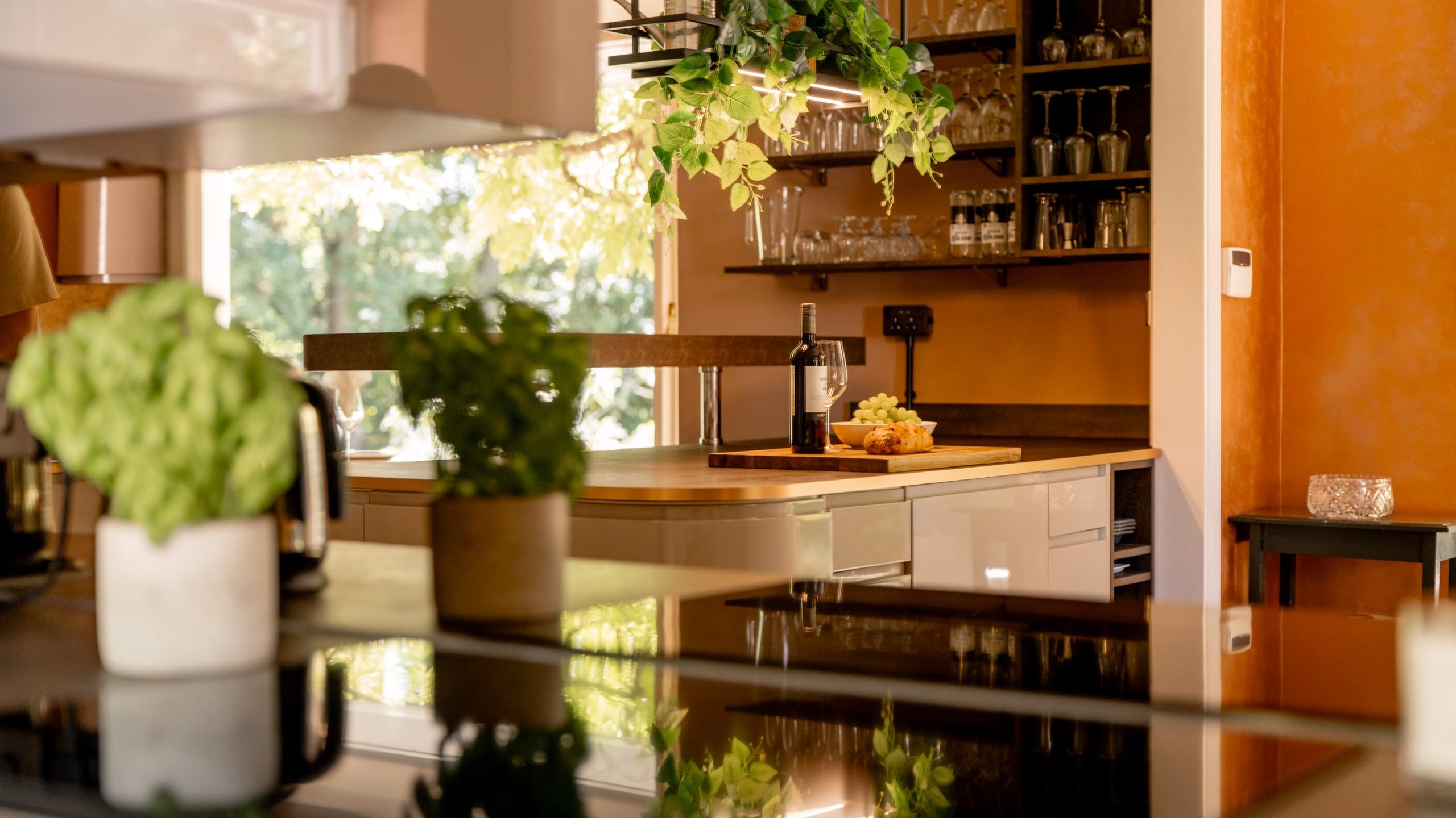 A modern kitchen with warm orange walls, hanging green plants, and shelves with glassware. Fresh herbs sit on the counter, and a bottle of wine with a cheese platter is displayed on a wooden island.
