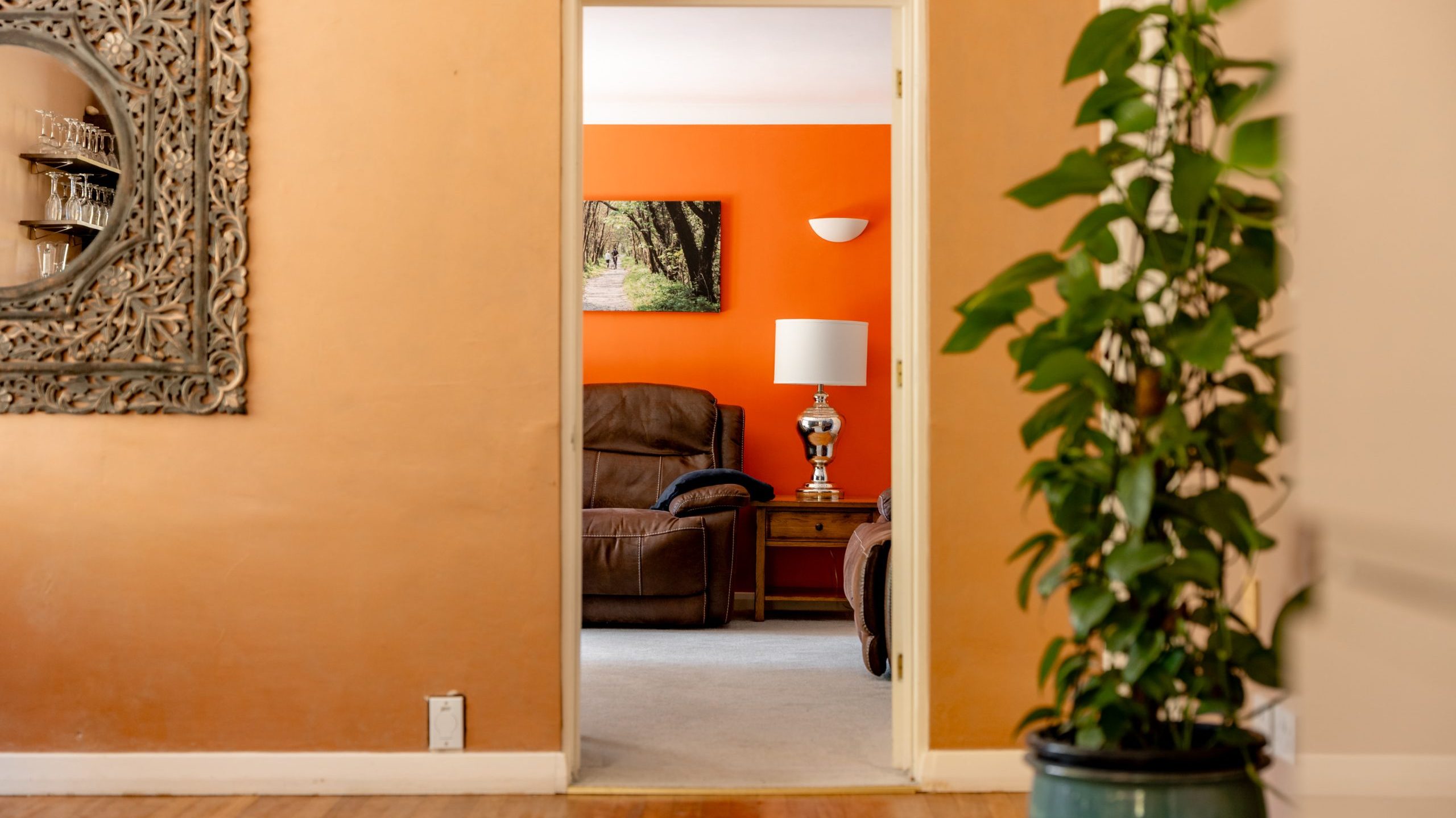 A view through a doorway shows a cozy living room with orange walls, a brown recliner, a table lamp, and a framed photo. A large green potted plant and a decorative mirror are seen in the foreground.