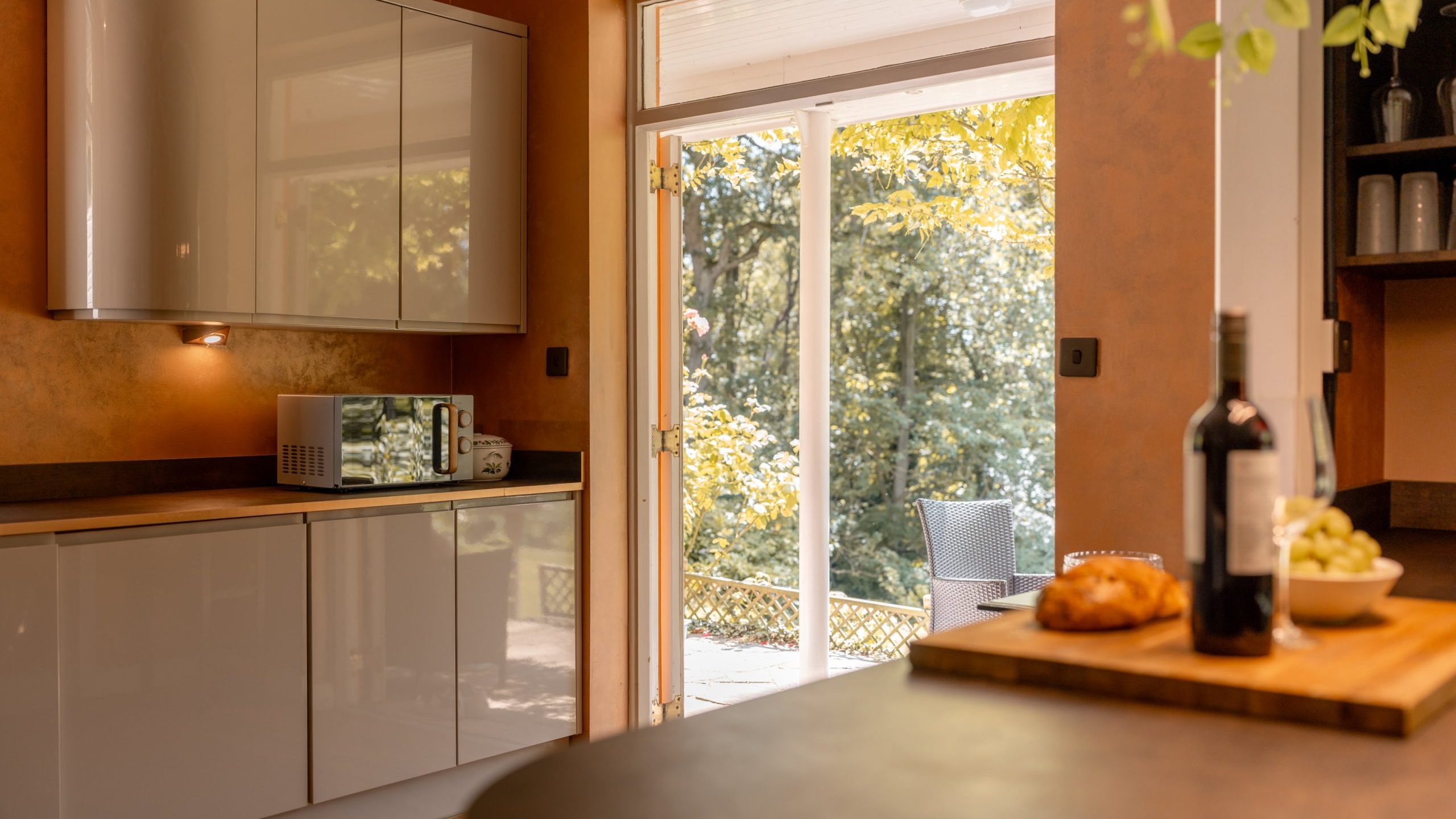 Modern kitchen with glossy white cabinets and a microwave on the counter, brightened by natural light from glass doors opening to a patio with chairs and lush greenery outside. A wine bottle, bread, and grapes are on the counter.