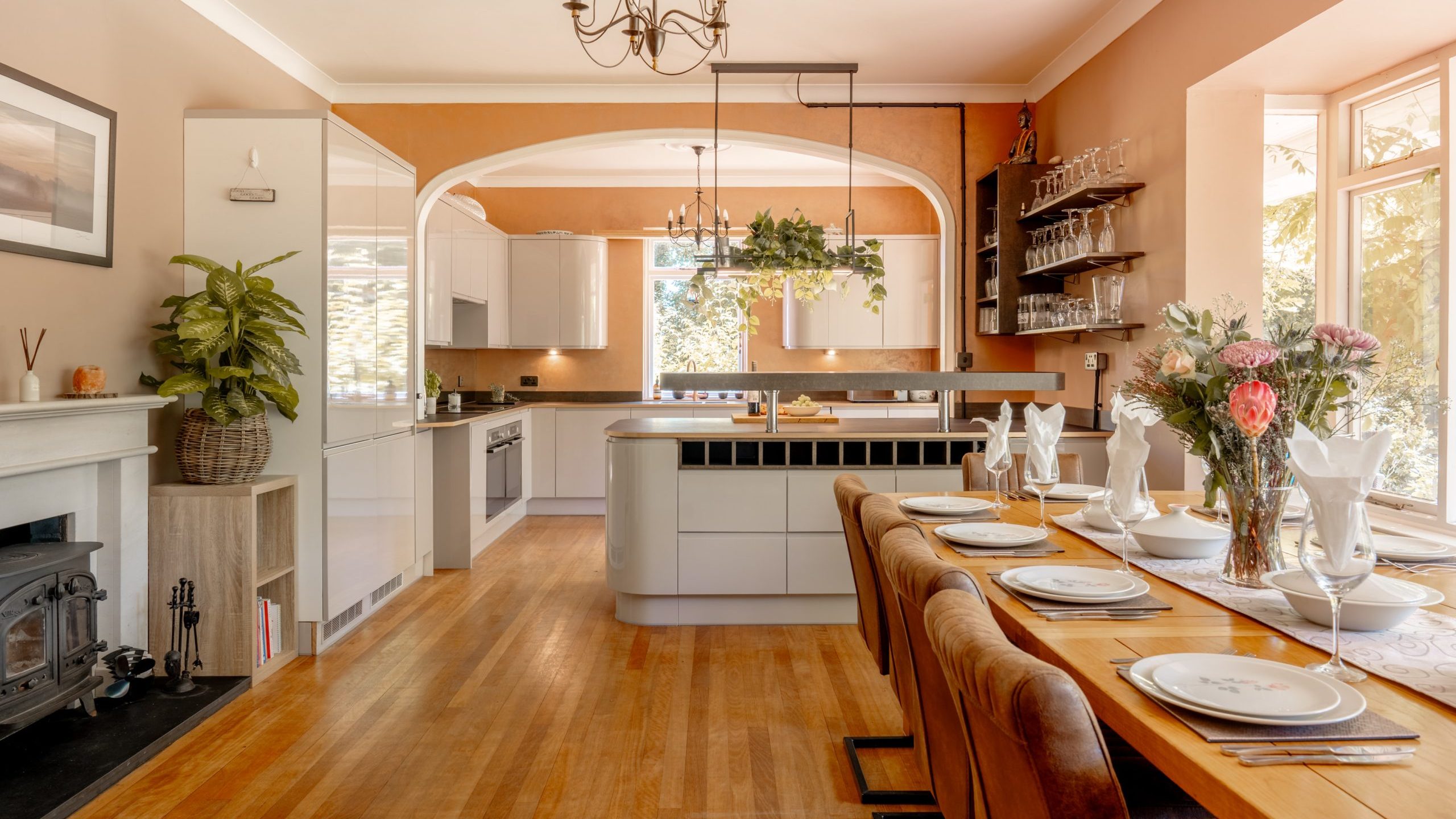 Bright, open-plan kitchen and dining area with wooden floors, a modern kitchen island, brown chairs at a set dining table, large windows, potted plants, shelves with glassware, and warm, neutral walls.