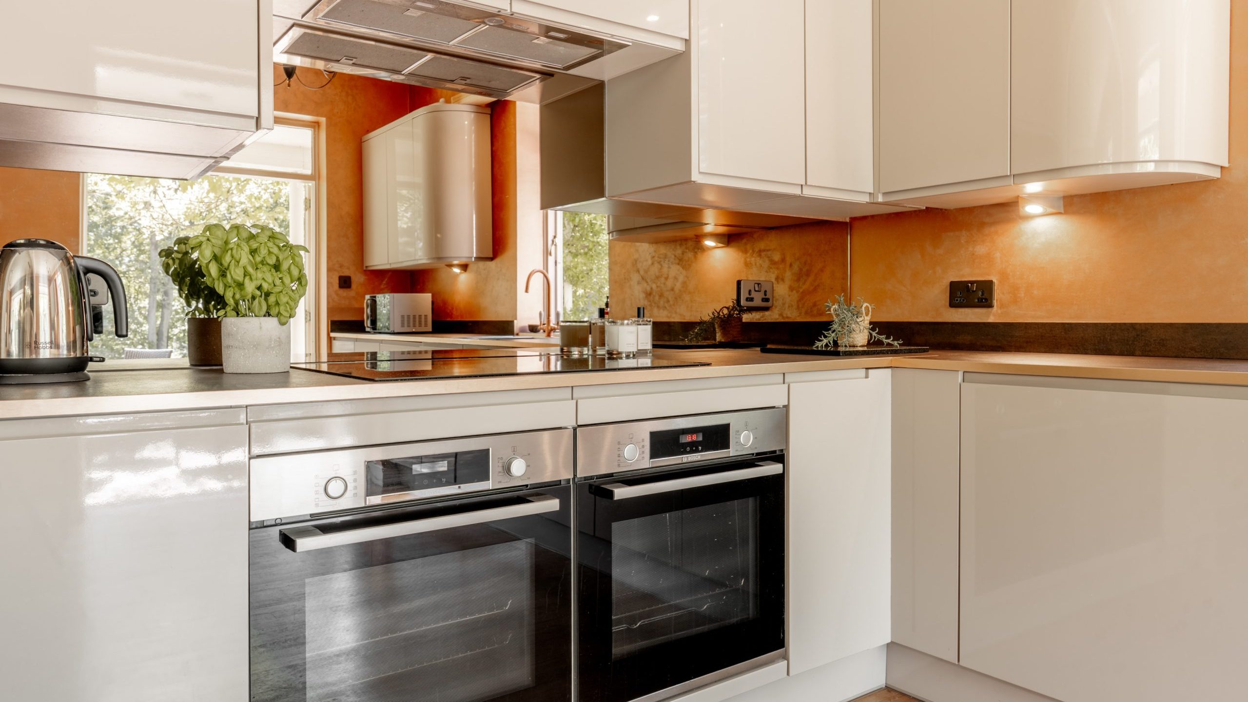 Modern kitchen with glossy white cabinets, built-in double oven, electric stovetop, stainless steel kettle, and potted basil plant. Warm orange walls and under-cabinet lighting create a cozy atmosphere.