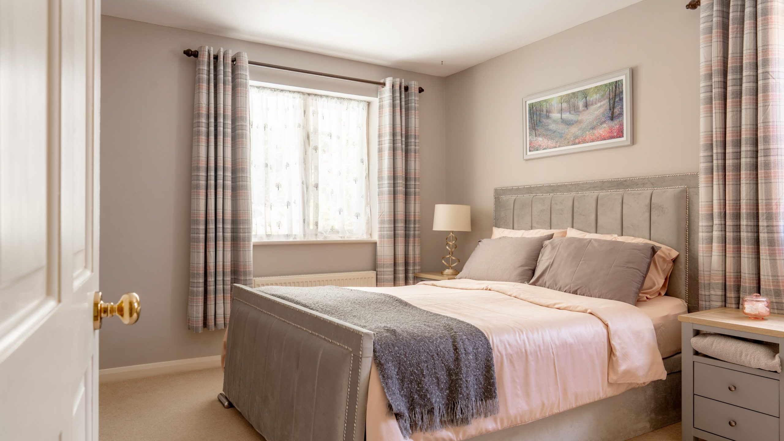 A cozy bedroom with a neatly made bed featuring pale pink bedding, gray headboard, and a gray throw. There are two windows with patterned curtains, a table lamp, and a framed landscape artwork above the bed.