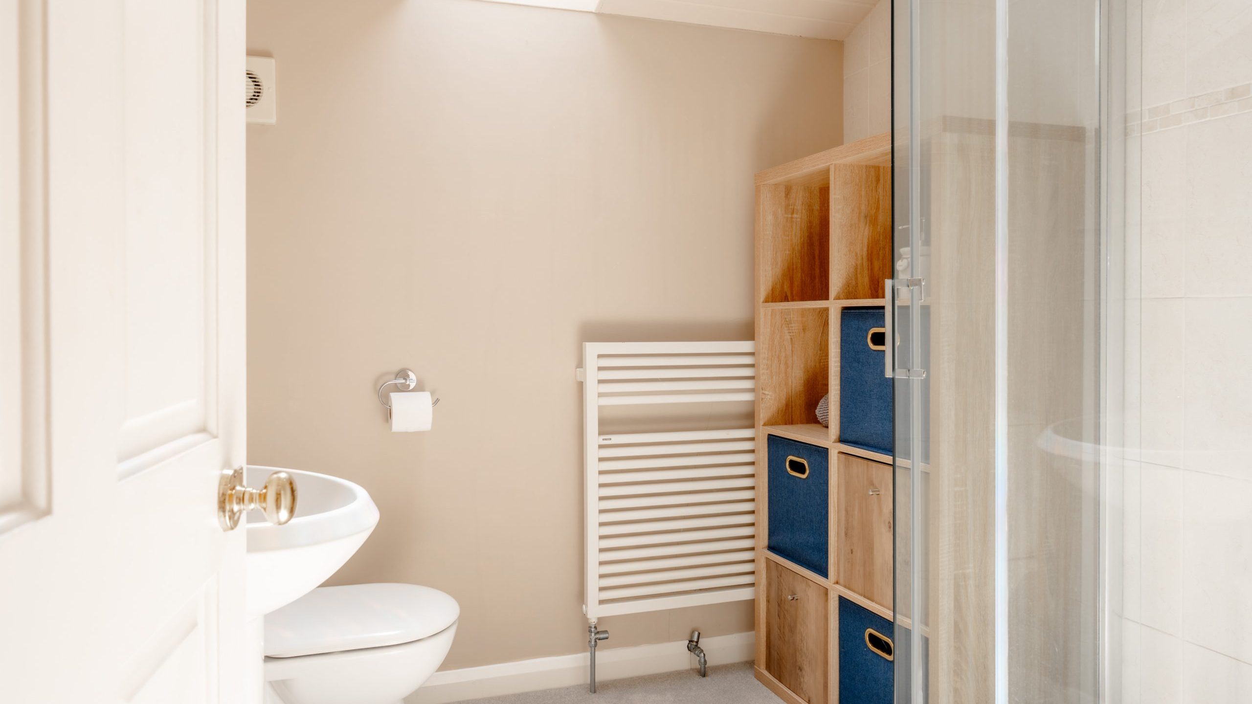 A modern bathroom with beige walls, a white toilet and sink, a glass shower enclosure, a white heated towel rail, and wooden shelves with blue storage baskets under a skylight.