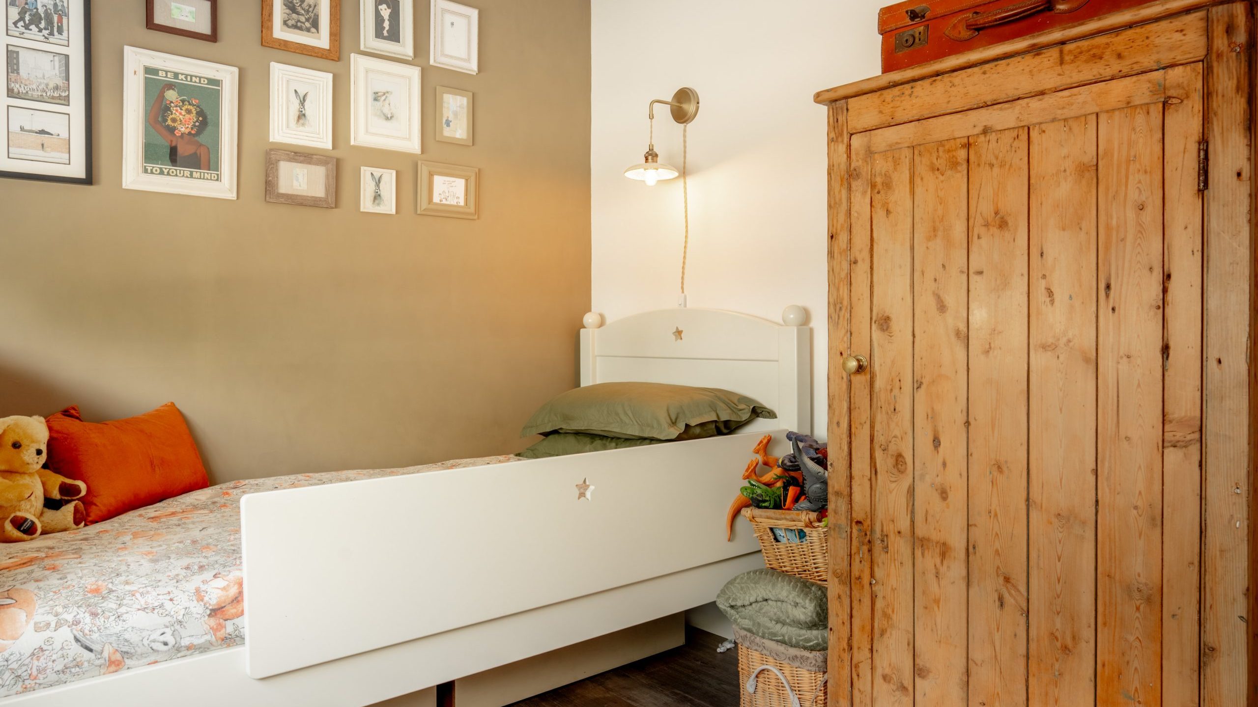 Cozy bedroom with a white bed, green pillows, teddy bears, a wicker basket of toys, a wooden wardrobe, and framed pictures on a beige wall under a wall-mounted lamp.