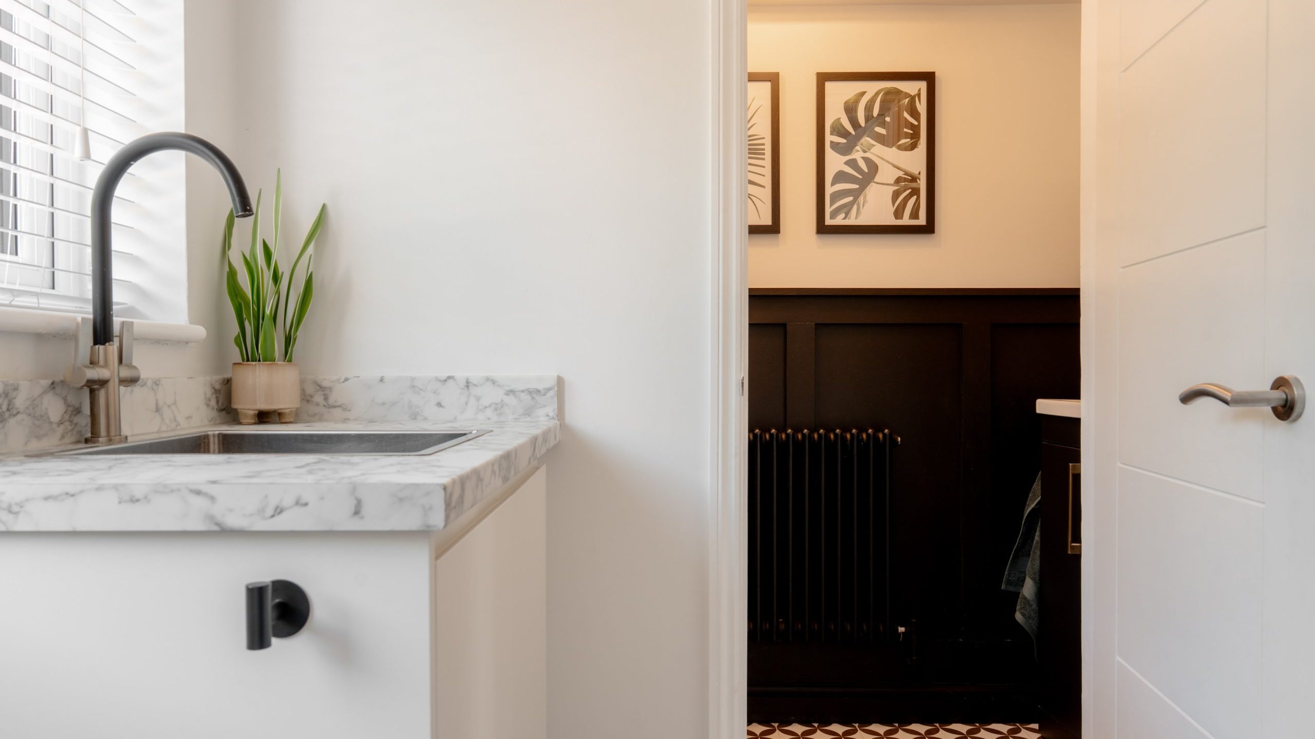 Modern bathroom with a marble countertop and sink, a potted plant, geometric floor tiles, and a view into an adjoining room with dark wall paneling and framed botanical artwork.