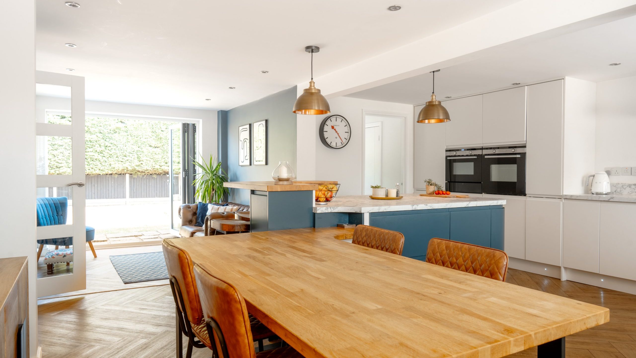 Modern kitchen and dining area with a wooden table and brown chairs, white cabinets, hanging pendant lights, a wall clock, and large windows letting in natural light. A seating area is visible in the background.