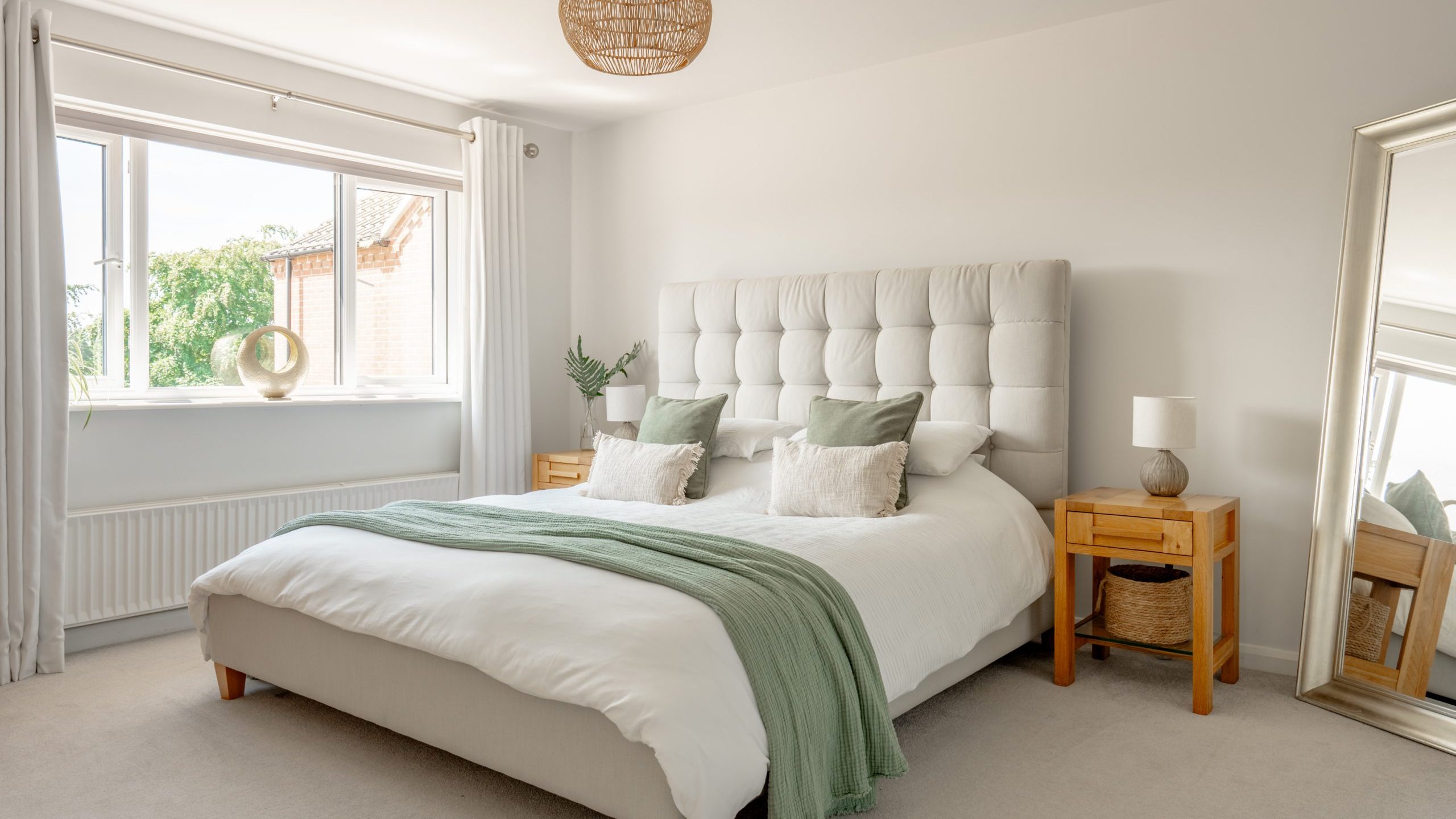 A bright, modern bedroom with a large bed featuring a tufted headboard, green throw blanket, and decorative pillows. Wooden nightstands with lamps and a large floor mirror sit beside the bed. Sunlight streams in through a window.