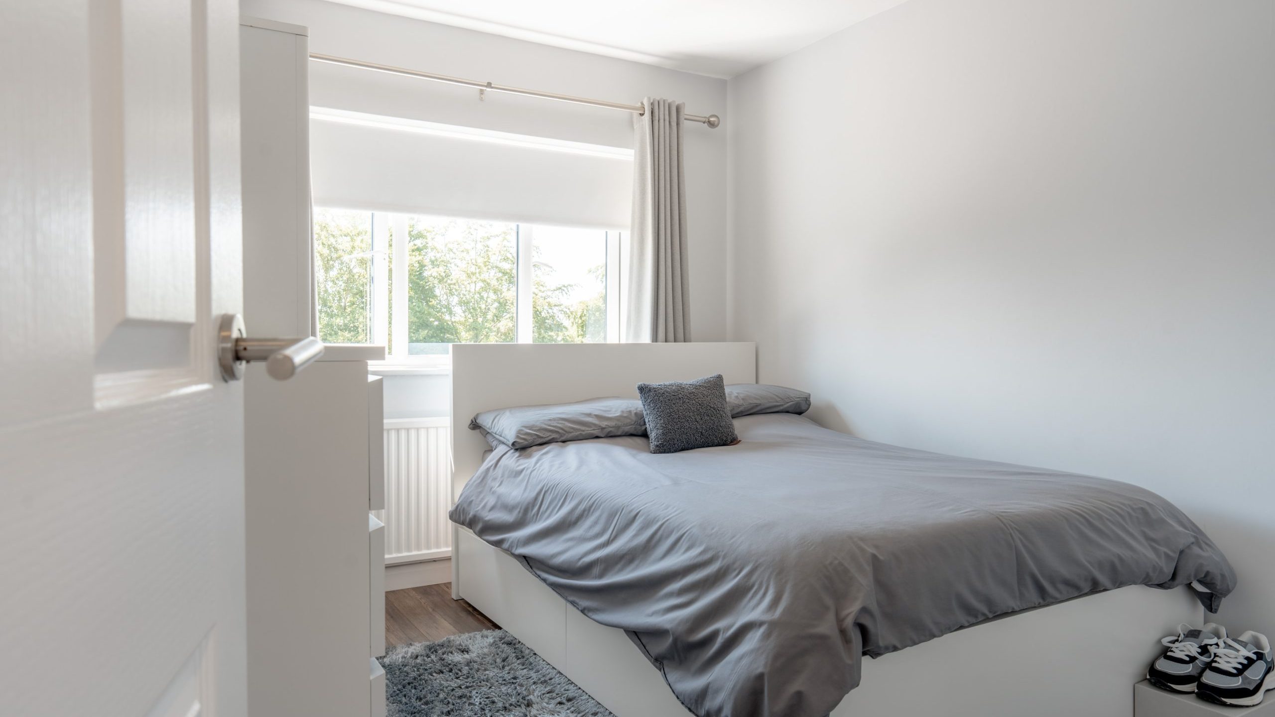 A bright, minimalist bedroom with a double bed covered in gray bedding, a small gray pillow, light gray curtains, a gray rug, and large windows letting in natural light. A pair of slippers is placed by the bed.