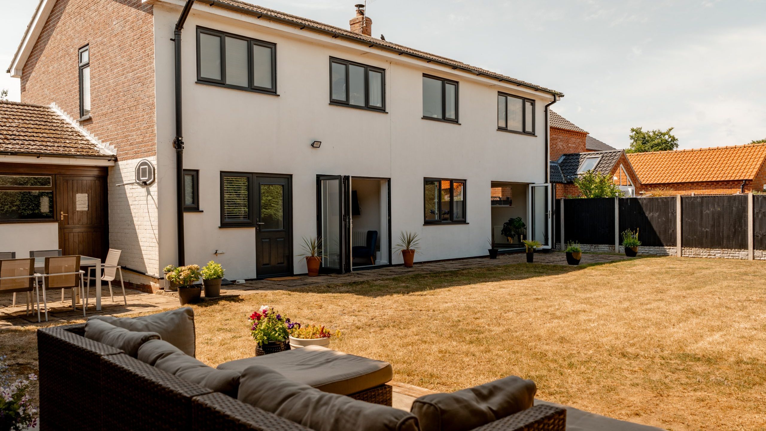 A modern two-story house with white walls and large windows, featuring a backyard patio with outdoor seating, potted plants, and a dry grassy lawn under a clear sky.