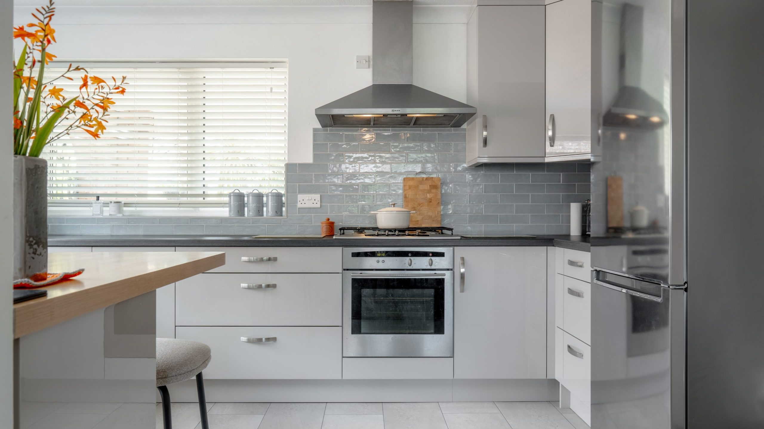 Modern kitchen with glossy gray cabinets, stainless steel appliances, a built-in oven, gas stove, and a fridge. A vase with orange flowers sits on the counter, and sunlight streams through white blinds.