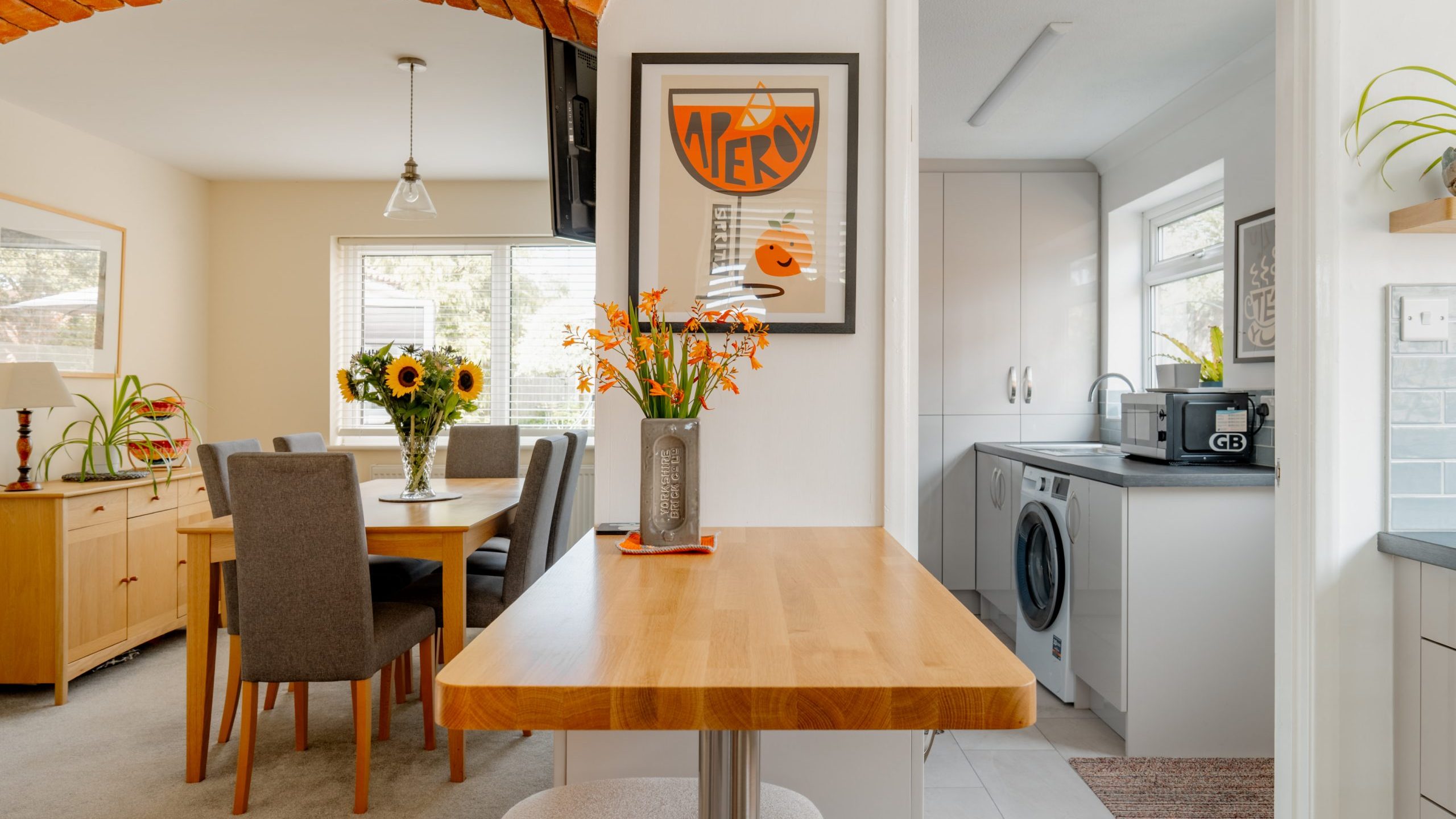 A cozy dining area with a wooden table, gray chairs, and sunflowers in a vase leads to a bright utility room with a washing machine, sink, and white cabinets. A framed orange art print decorates the wall.