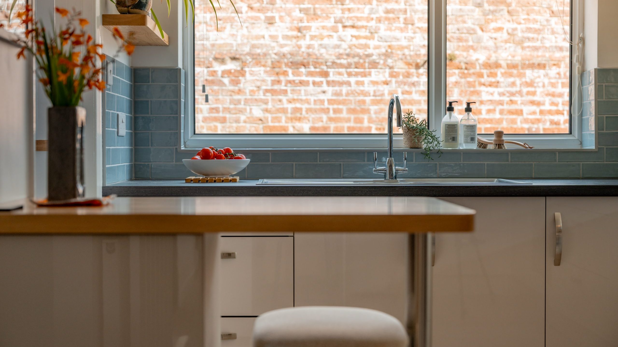 A modern kitchen with white cabinets, a light wood countertop, a stool, and a large window above the sink. A plant, soap, and a bowl of tomatoes sit on the counter. Blue subway tiles line the backsplash.