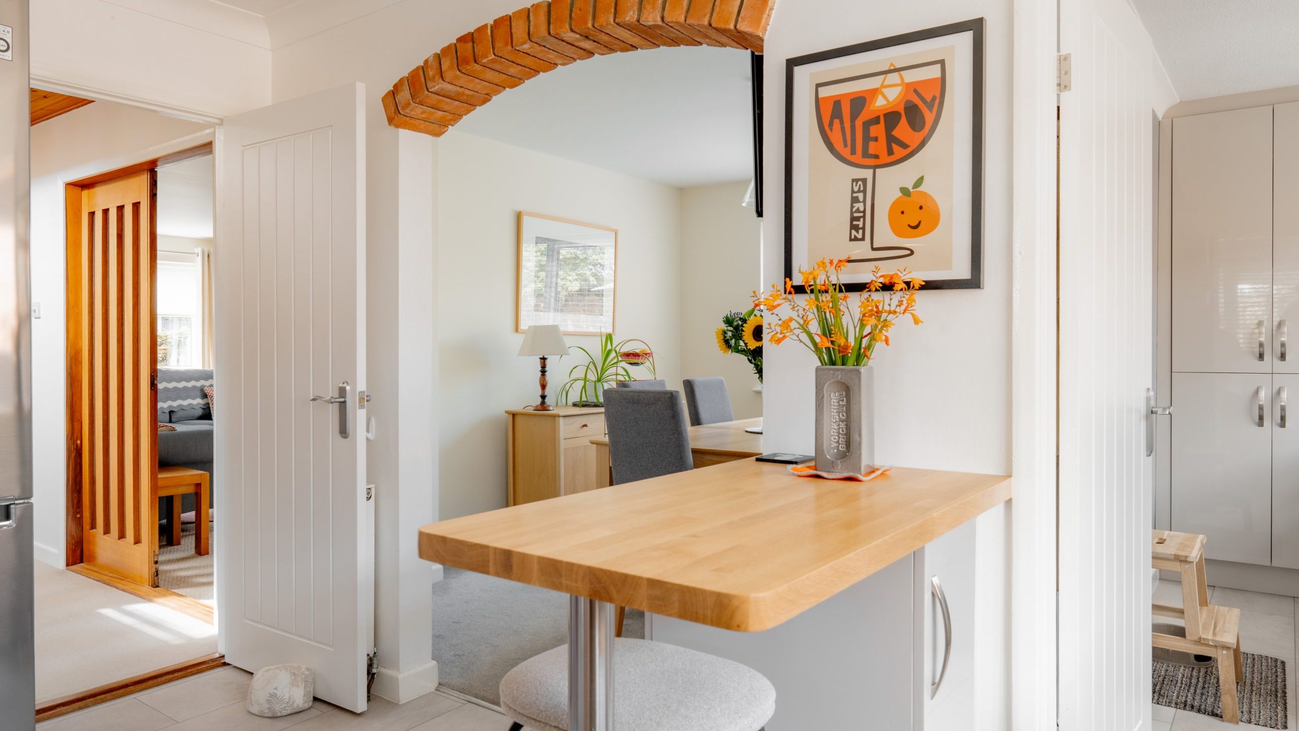 A bright kitchen with a wooden breakfast bar, stool, and vase of orange flowers. An arched brick doorway leads to a cozy living area with light walls, furniture, and framed art on the walls.