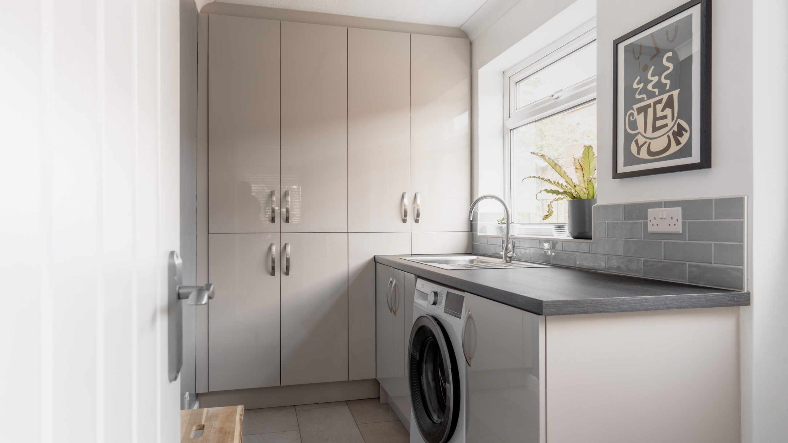 Modern laundry room with beige cabinets, a washing machine under a black countertop, a sink by a window with a potted plant, light gray tile backsplash, and framed artwork on the wall.