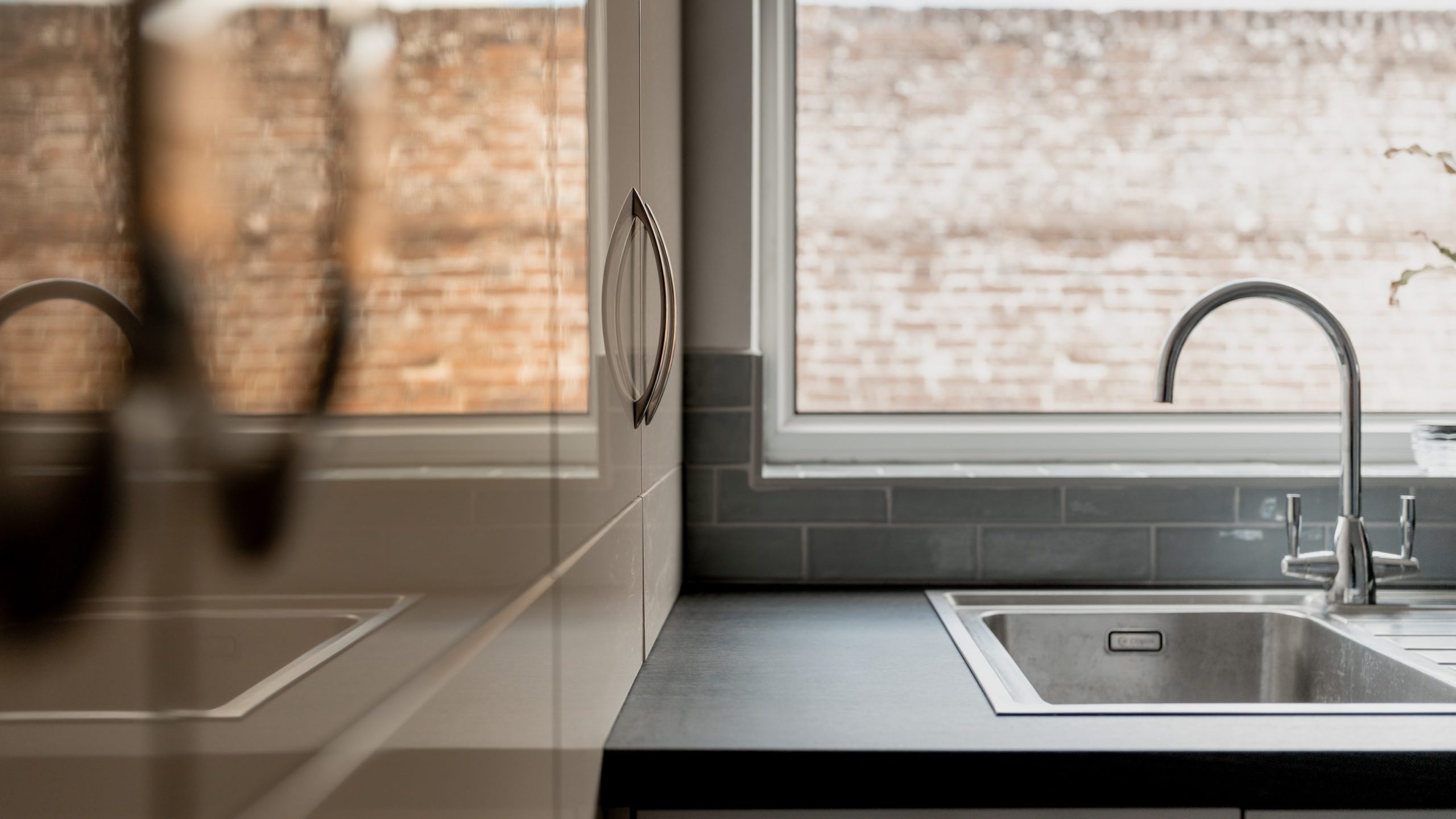 Modern kitchen with a sleek black countertop, stainless steel sink, chrome faucet, and a large window letting in natural light; reflection of the countertop is visible on the glossy cabinet surface.