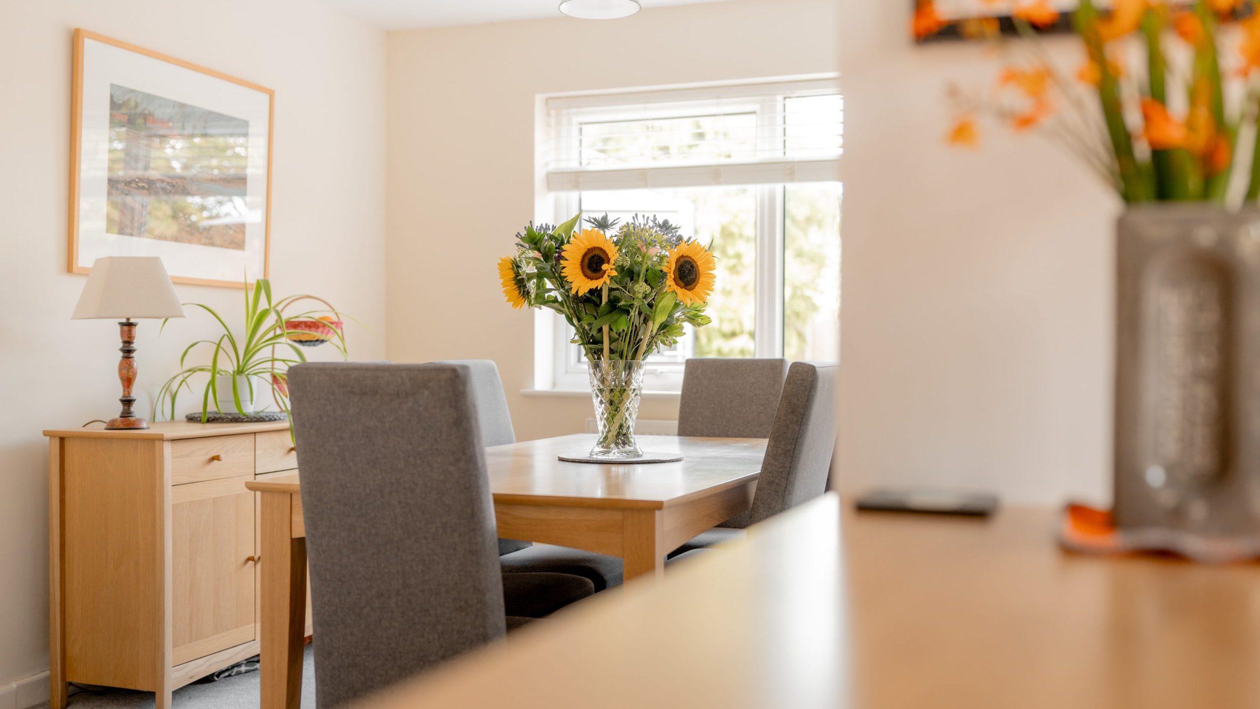 A bright dining room with a wooden table, four gray chairs, and a vase of sunflowers in the center. Sunlight streams through a window, and there are plants and framed art on light-colored walls.
