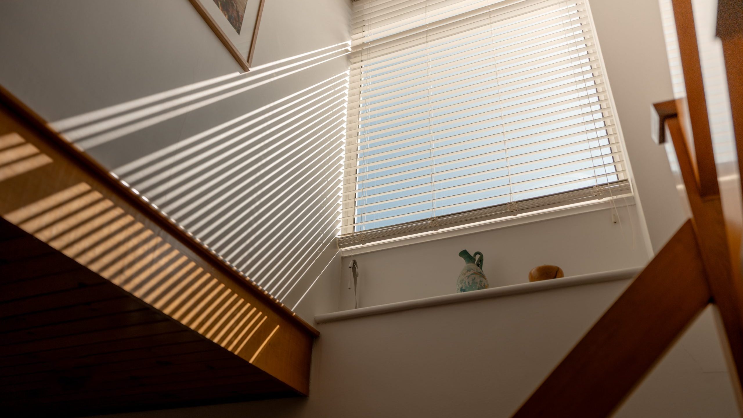 Sunlight streams through white blinds onto a stairwell, casting striped shadows on the wall and stairs. A framed picture hangs on the wall, and two decorative objects sit on the windowsill.