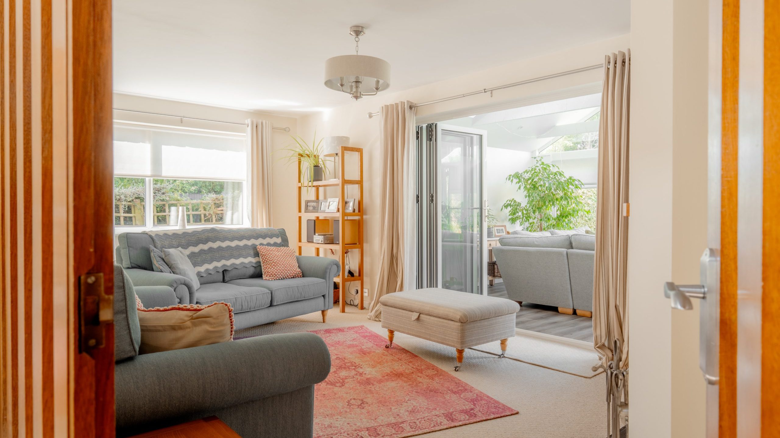 Bright living room with grey sofas, a pink rug, a wooden bookshelf, and a large ottoman. Glass doors open to a sunroom with more seating and a large potted plant. Natural light fills the space.