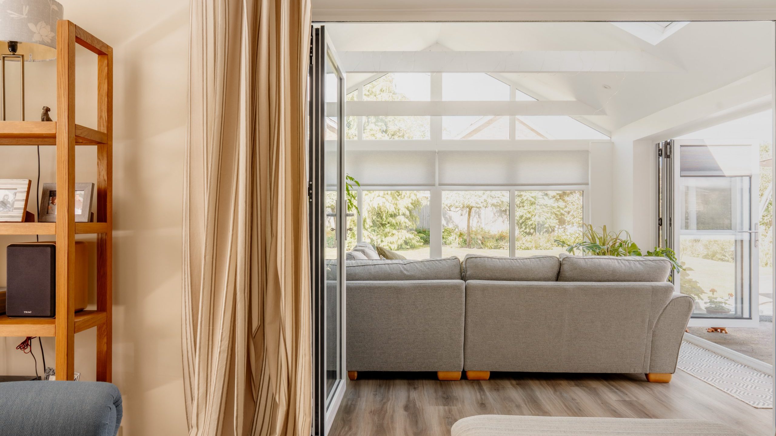 A bright living room with a large L-shaped gray sofa near wide windows, letting in natural light. Beige curtains and a wooden shelf are visible in the foreground. The space appears open and airy.