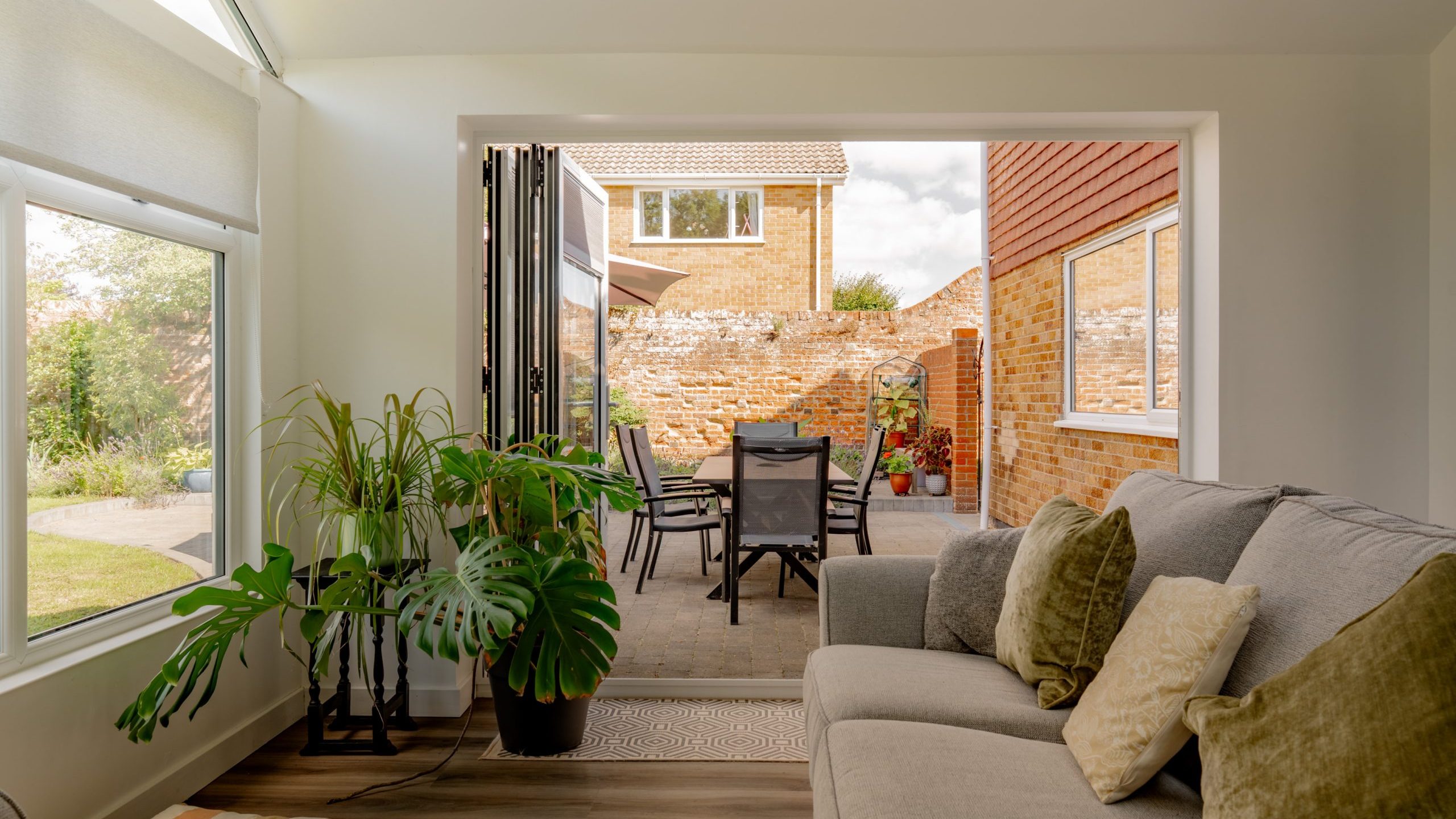 A cozy living room with a gray sofa and indoor plants opens onto a patio with a dining table and chairs, surrounded by brick walls and a garden visible through large glass doors.