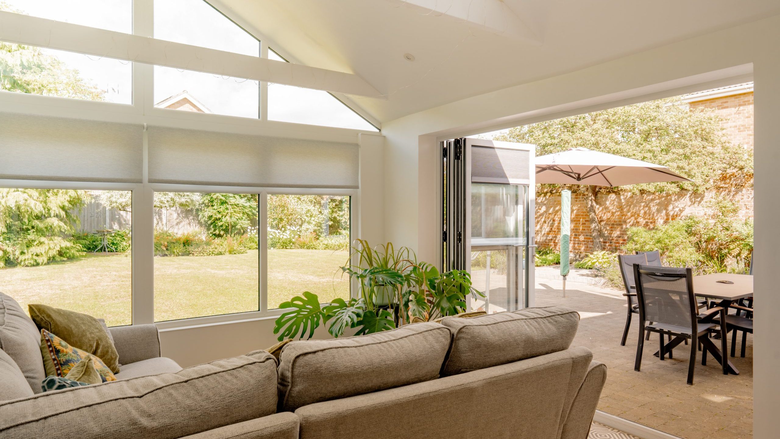 A bright sunroom with large windows and a gray sofa overlooks a patio with a table, chairs, and a beige umbrella, opening to a green garden with trees and shrubs.