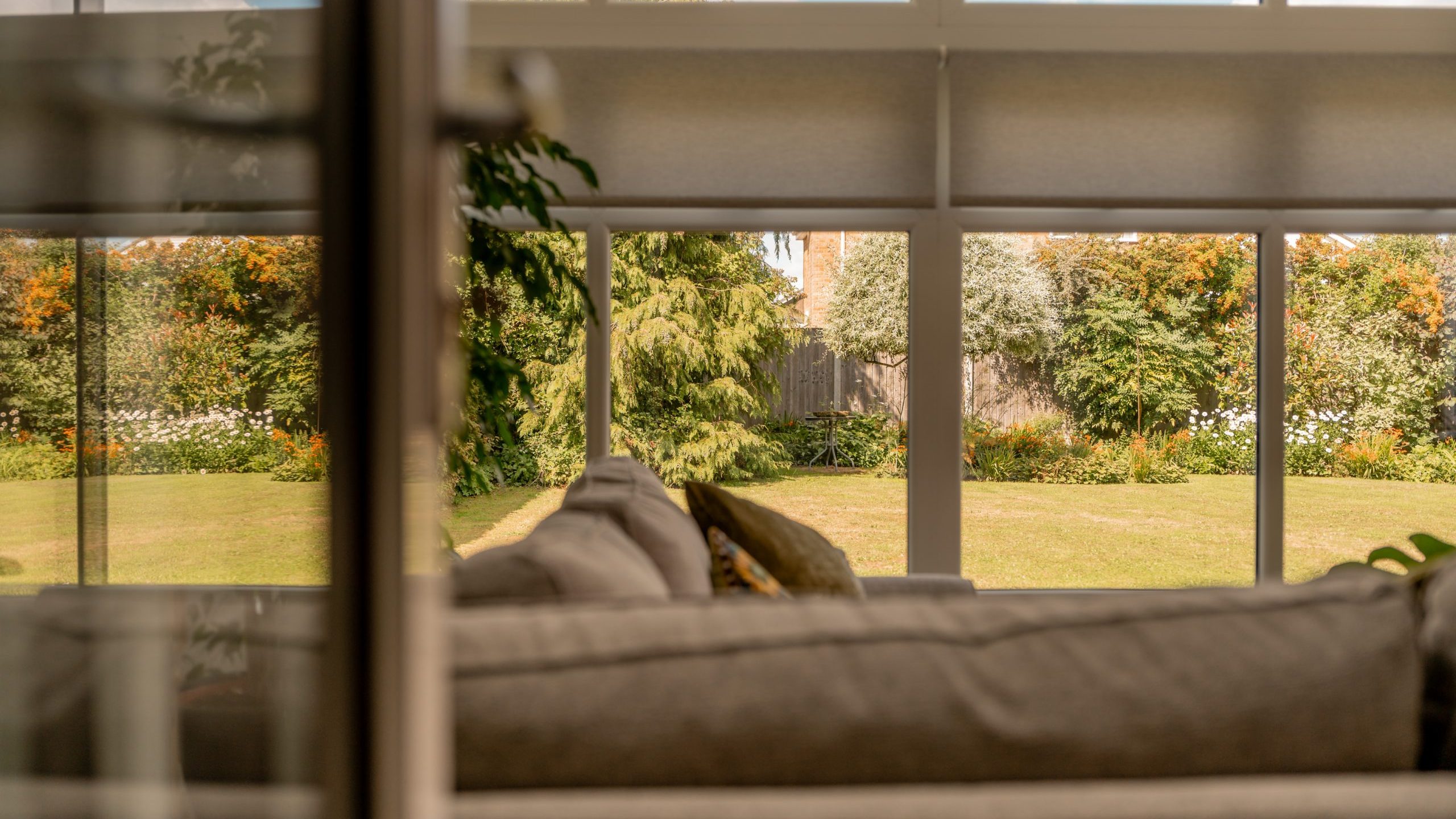 A cozy living room with a beige sofa faces large windows, offering a clear view of a green garden and trees outside on a sunny day.