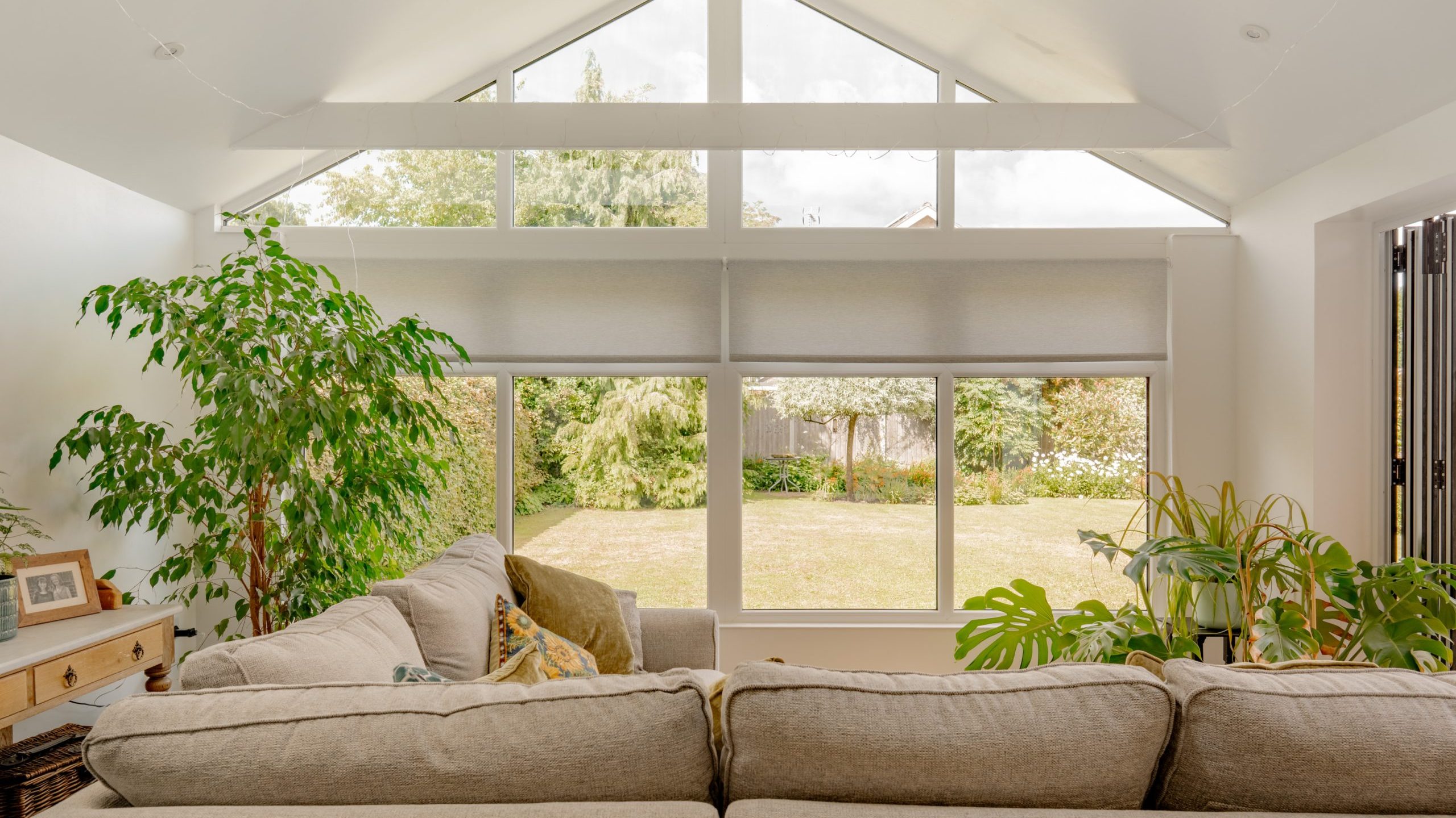 A bright living room with large windows overlooking a garden, beige sofas, green indoor plants, a wooden side table with a photo frame, and high, sloped ceilings with exposed beams.
