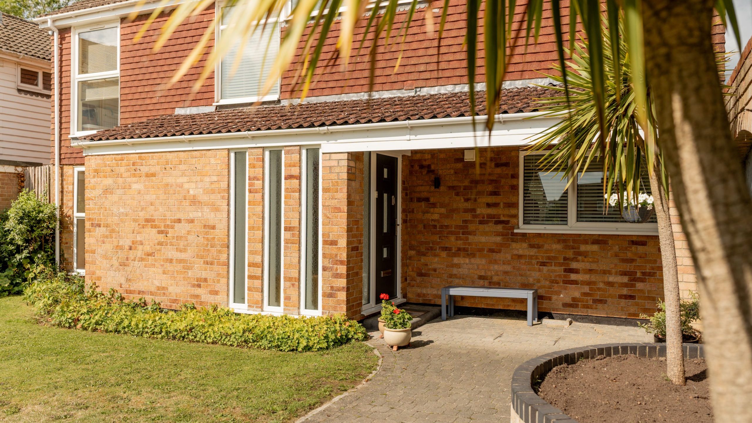 A two-story brick house with a front garden, a paved walkway leading to the entrance, tall windows, a small bench by the door, and a palm tree in the foreground.