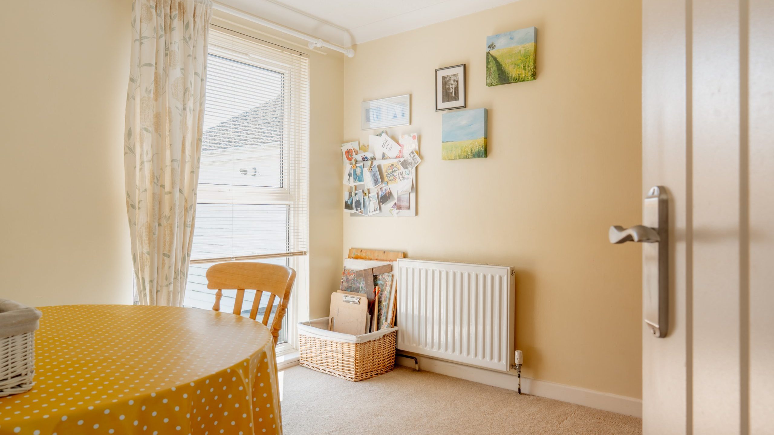 A bright room with beige walls, a window with sheer curtains, a table with a yellow polka-dot tablecloth, a wooden chair, a white radiator, and framed pictures and artwork on the wall above a basket.