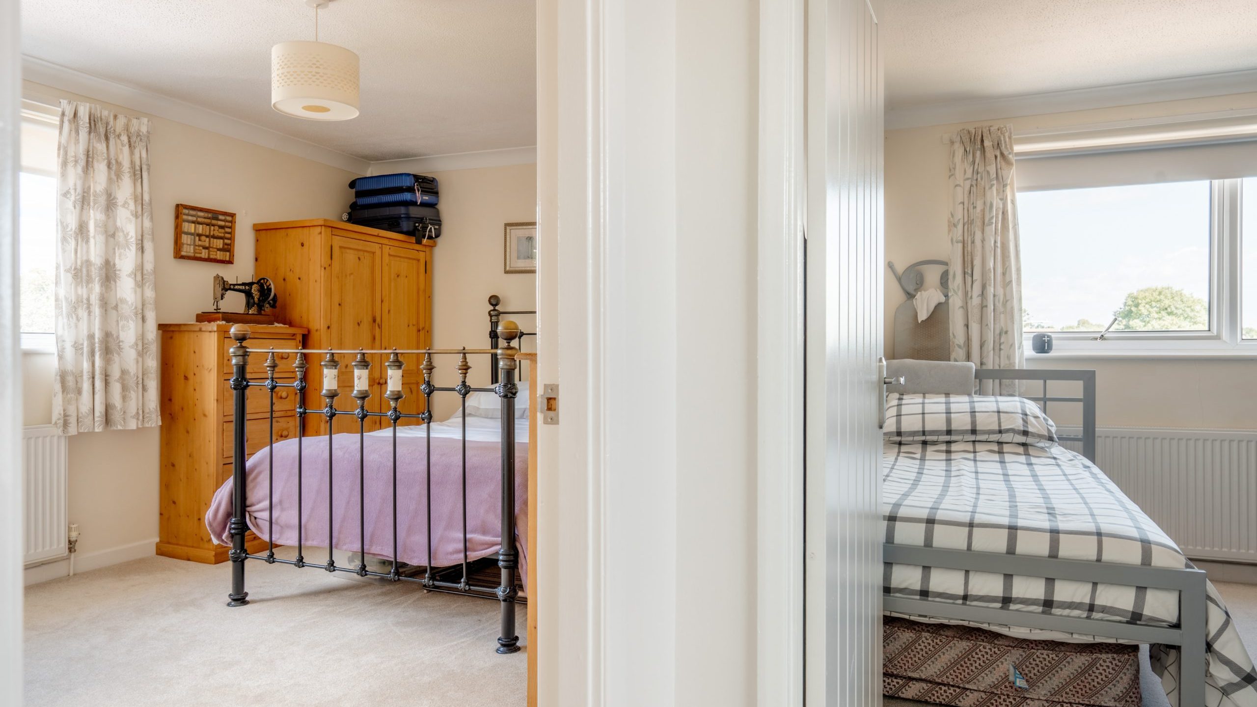 A view from a hallway showing two bedrooms: one with a metal bed frame, wooden wardrobe, and purple bedding; the other with a single bed, checkered bedding, and a large window with natural light.