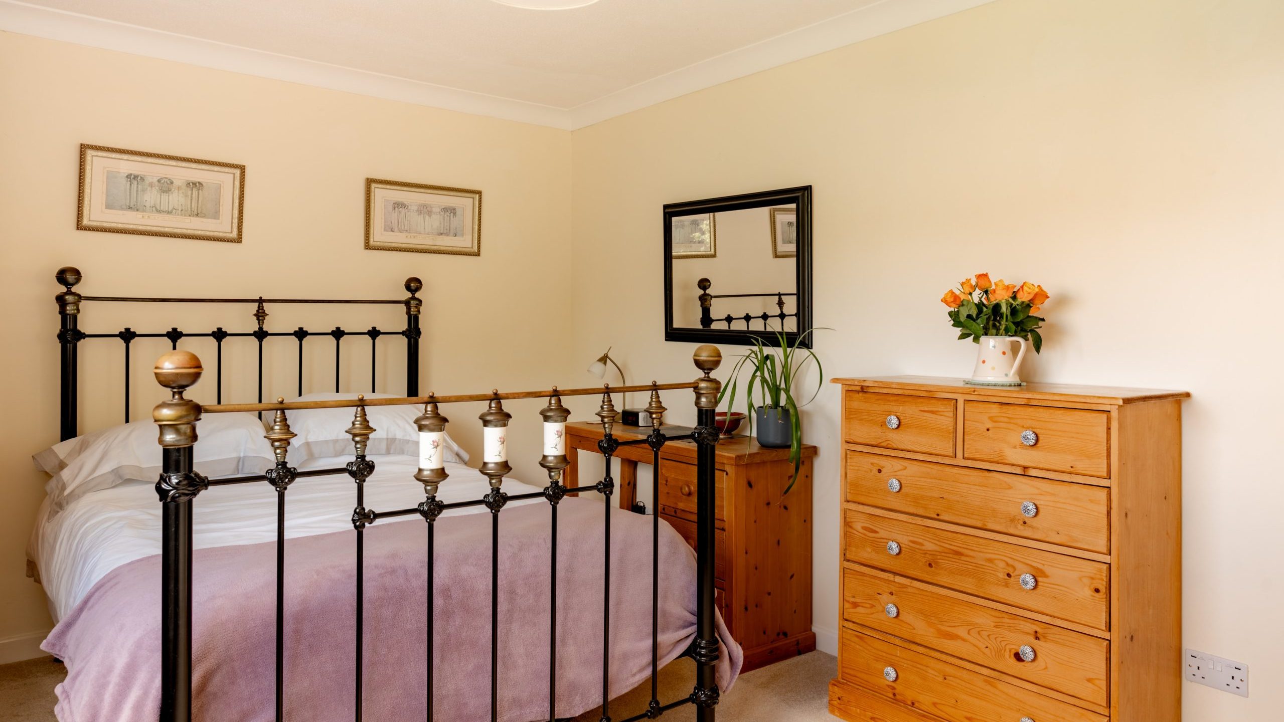 A cozy bedroom with a black metal bed frame, white bedding, and a pale purple blanket. There is a wooden dresser with orange flowers in a vase, a matching nightstand, a mirror, and framed art on cream-colored walls.