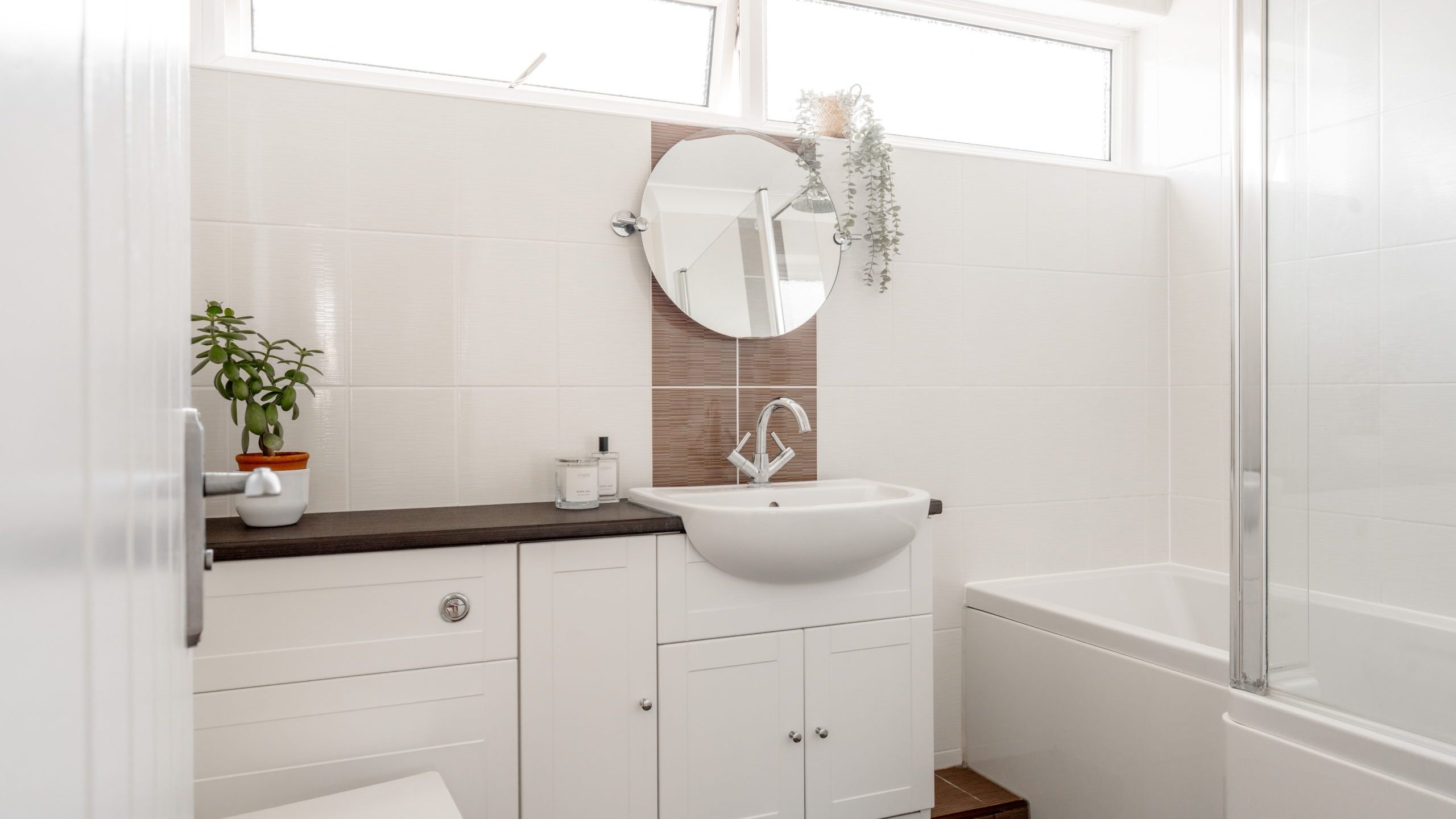 A bright, modern bathroom with white cabinets, a round mirror above a sink, potted plants on the counter, and a bathtub with a glass screen. Natural light enters through two high windows.