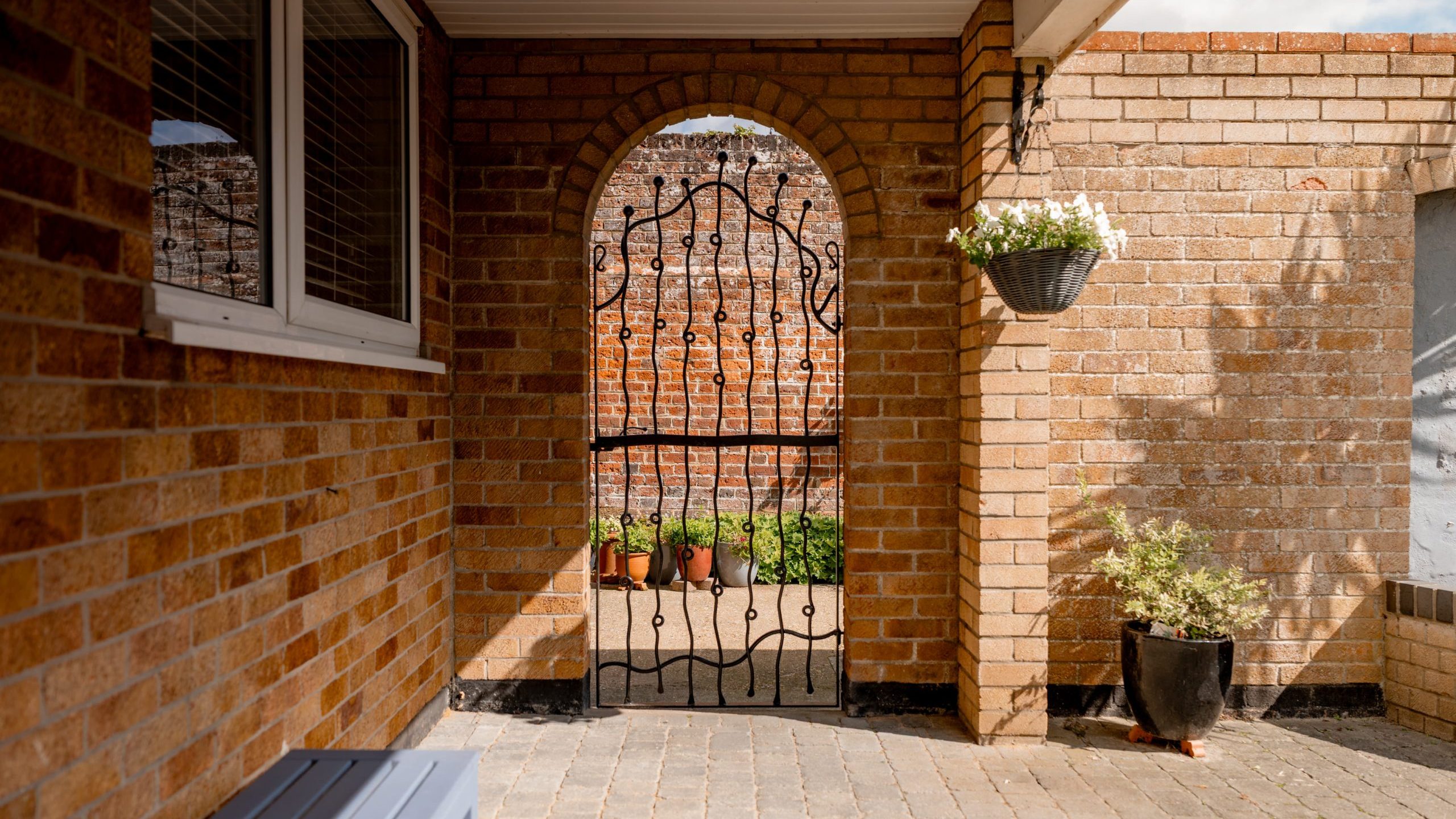 A brick patio area with a metal gate set in an arched brick opening. There are potted plants beyond the gate, a hanging basket with flowers, a potted plant on the right, and a bench on the left.