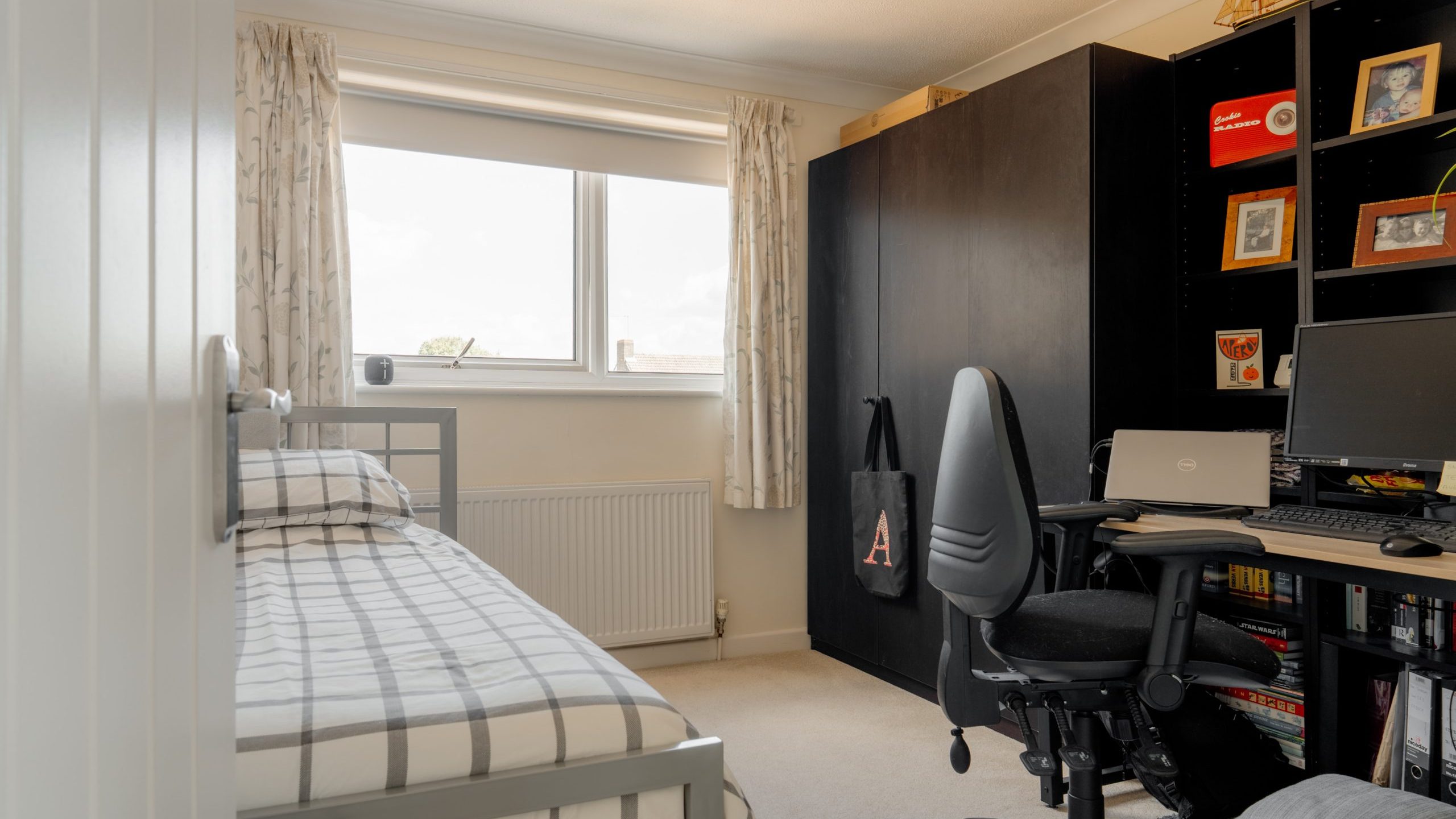 A tidy bedroom with a single bed, plaid bedding, a large black wardrobe, a desk with dual monitors, office chair, bookshelves, and a window with curtains letting in natural light.