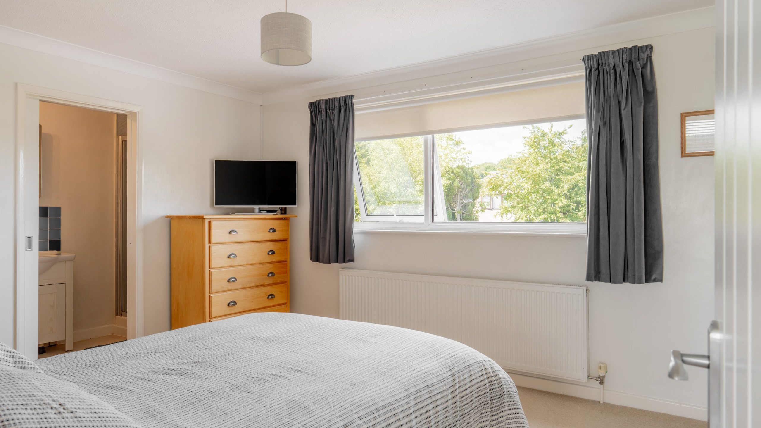 Bright bedroom with a double bed, gray bedding, wooden dresser, mounted TV, large window with gray curtains, and an open door leading to a bathroom. Walls are white, and natural light fills the room.