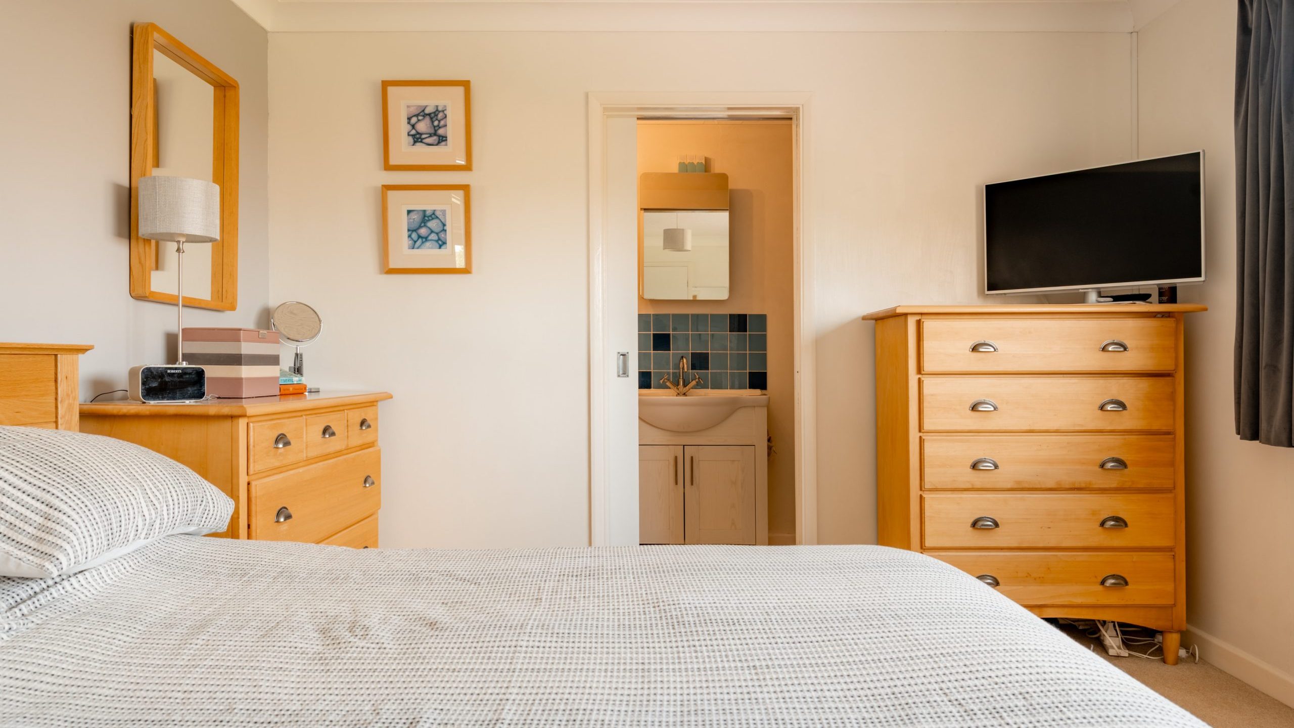 A cozy bedroom with light wood furniture, including a bed, dresser, and chest of drawers. A flat-screen TV sits on the chest, and an en-suite bathroom with a sink and mirror is visible through an open door.