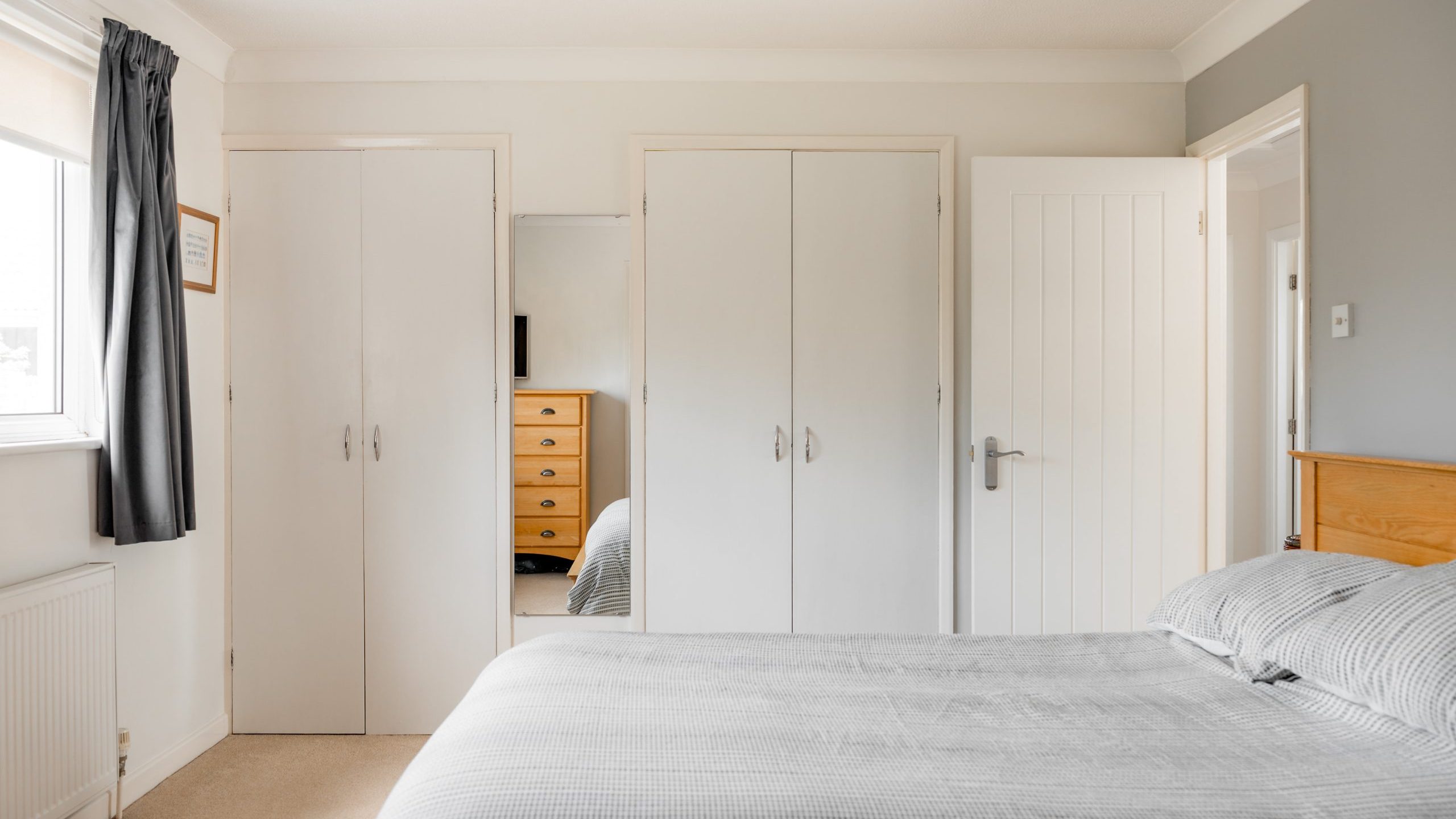 A bright bedroom with a double bed covered in gray striped bedding, fitted wardrobes with white doors, a wooden chest of drawers, and a window with gray curtains letting in natural light.