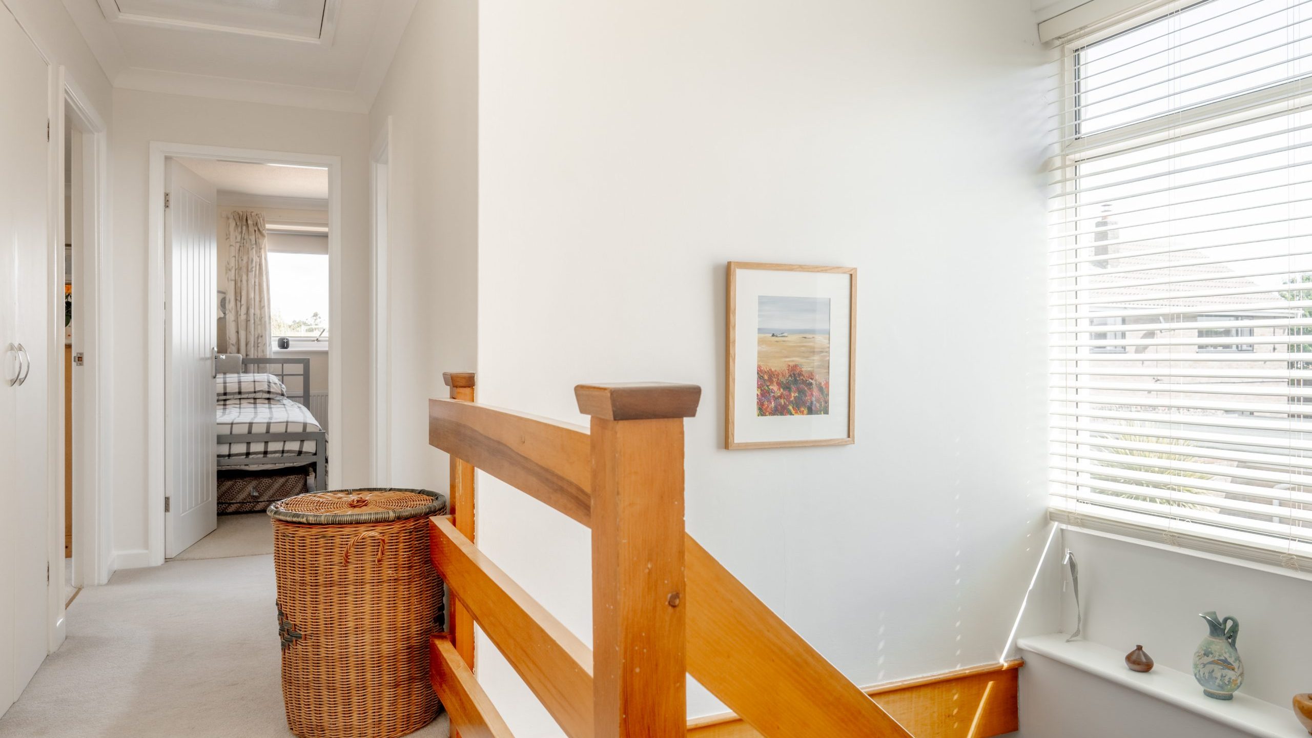 A bright, carpeted hallway with a wooden stair railing, a wicker laundry basket, a framed landscape picture on the wall, and a window with blinds letting in natural light. A bedroom is visible at the end of the hall.