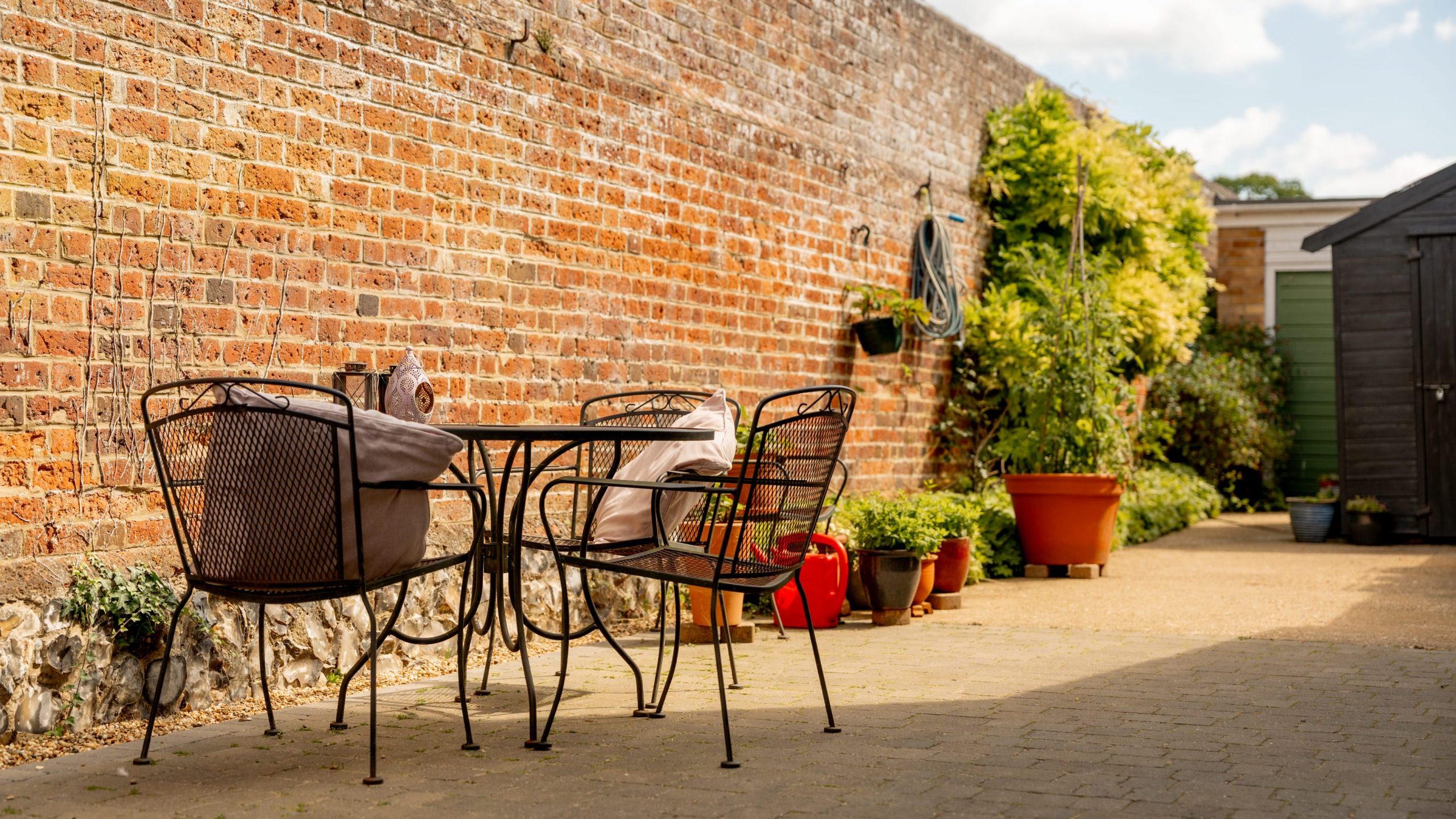 A sunny patio with a black metal table and four chairs, set against a tall brick wall. Potted plants and a garden hose decorate the area, and a dark shed stands in the background under a blue sky with clouds.