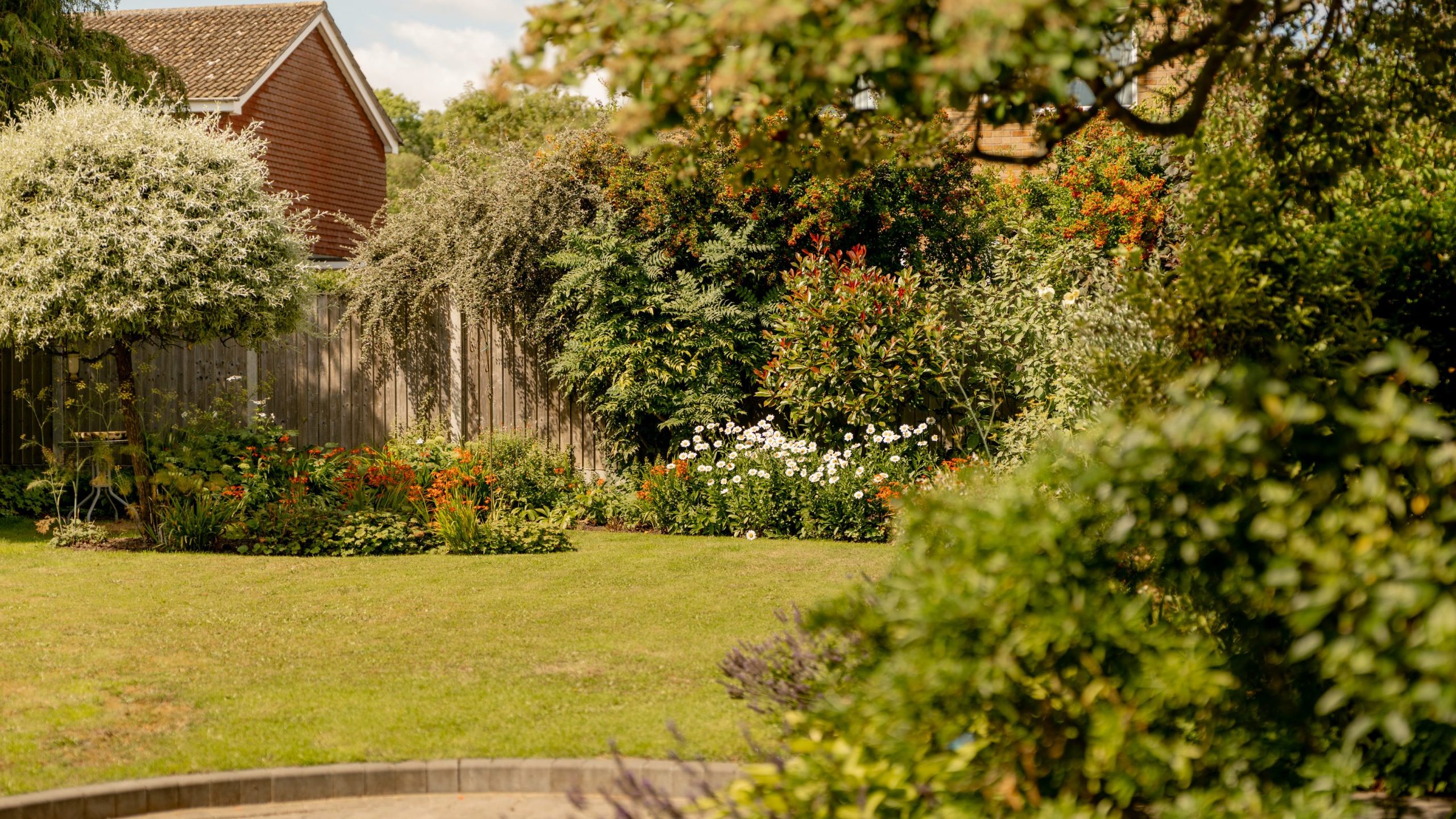 A lush garden with a green lawn, various bushes, colorful flowers, and a wooden fence, with a brick house partially visible in the background under a blue sky.