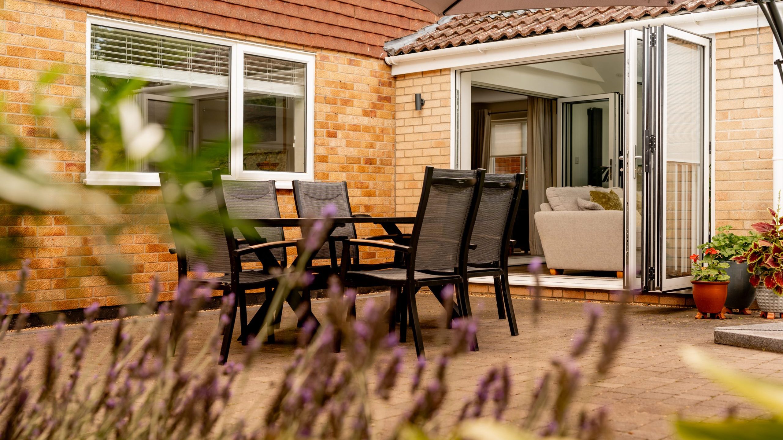 A modern patio with a black outdoor dining set and open glass doors leading to a cozy living room. Potted plants and purple flowers line the brick patio next to a beige brick house under a large umbrella.