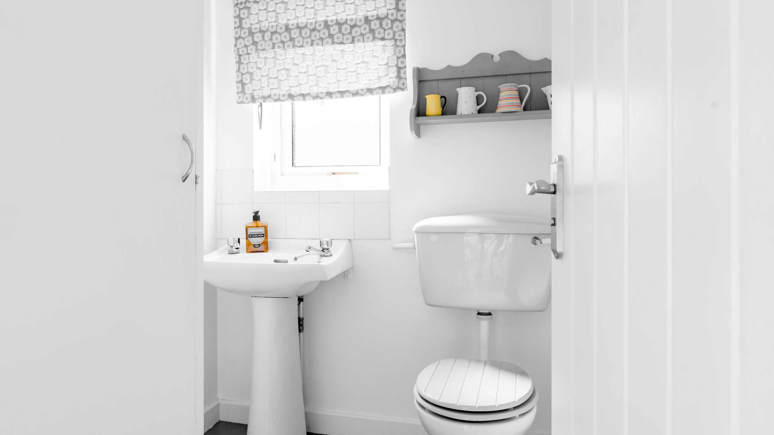 A small, bright bathroom with a white pedestal sink, a white toilet, shelf with cups above, a window with a patterned blind, and hand soap on the sink. The room has white walls and dark floor tiles.