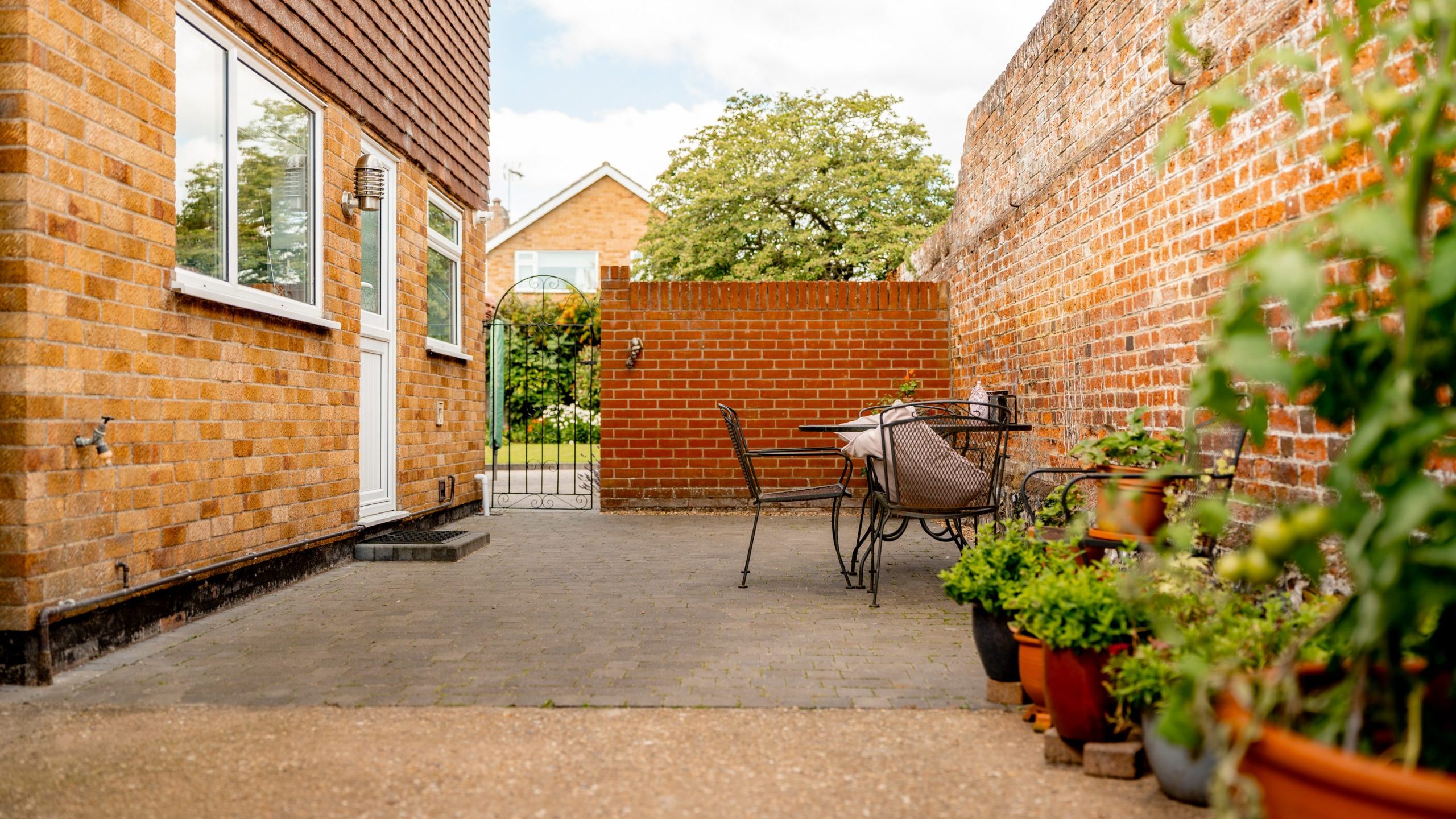 A brick patio with potted plants, a metal table and chairs, and a brick wall. A gate leads to a garden area, and a house with a window is visible on the left. The scene is bright and sunny.