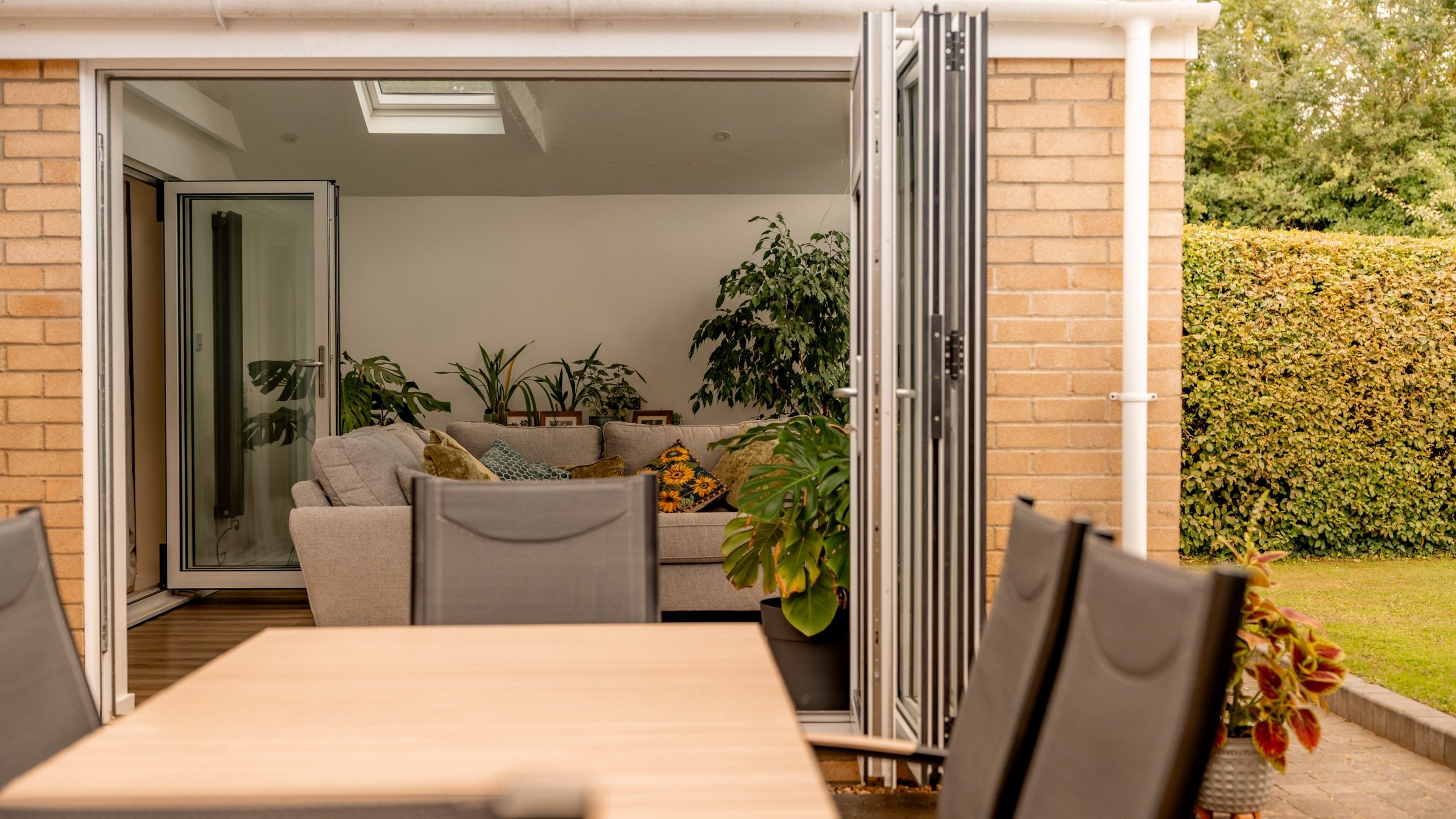 A modern patio with a wooden dining table and black chairs leads to a living room through open folding glass doors. Inside, there's a gray sectional sofa and several green houseplants. A hedge and lawn are visible outside.