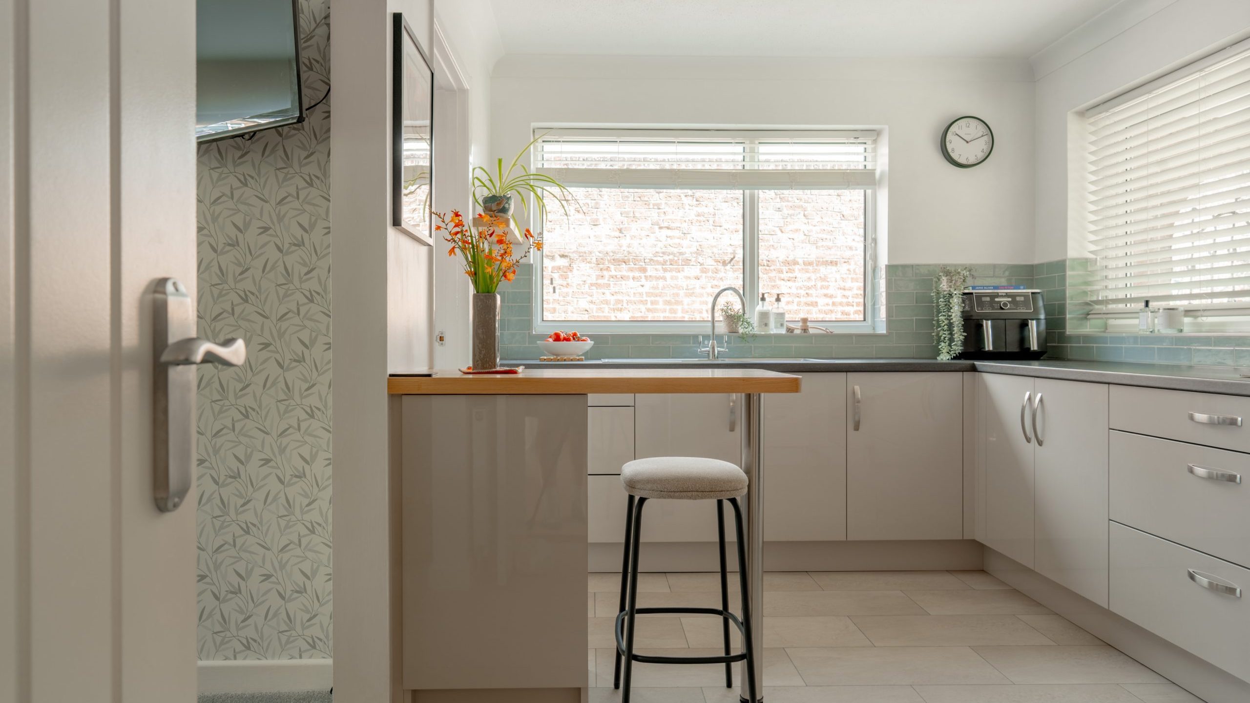 Bright, modern kitchen with white cabinets, light gray countertops, a high stool by the breakfast bar, green-tiled backsplash, sink under a large window, coffee maker, clock, and vase with flowers on the counter.