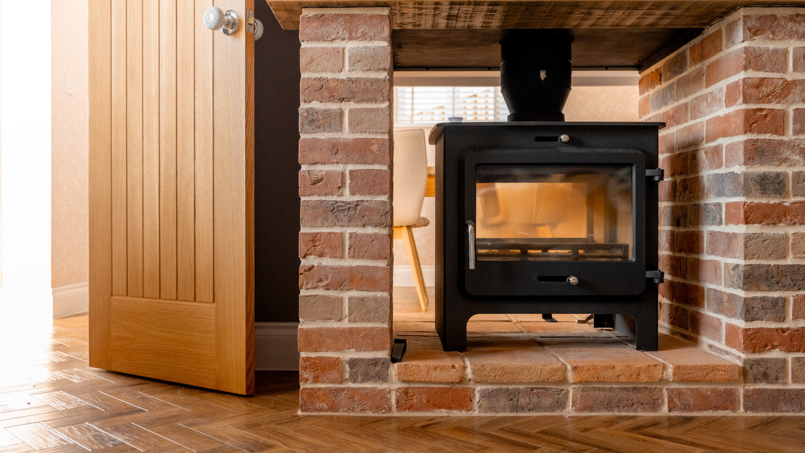 A black wood-burning stove set in a brick fireplace, with a wooden mantel above. An open wooden door and part of a dining table with chairs are visible in the background.